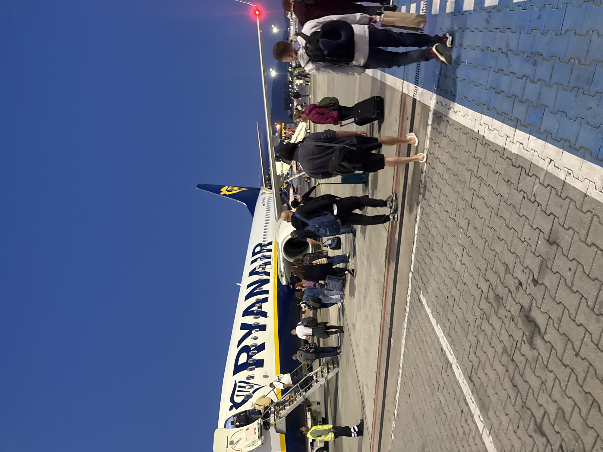 An evening scene at Sofia Airport, Bulgaria, shows passengers boarding a Ryanair airplane. The airplane, a Boeing 737, is positioned on the tarmac with its boarding stairs extended. Several individuals are visible, some ascending the stairs to board the aircraft, while others are waiting their turn or walking on the tarmac. Luggage, including suitcases and backpacks, is present with the passengers. A ground crew member in a high-visibility vest stands near the stairs, overseeing the boarding process. The sky is dark blue, indicating either early morning or late evening. The tarmac is paved with interlocking concrete blocks, marked with white lines. The overall environment suggests a typical airport departure scene.