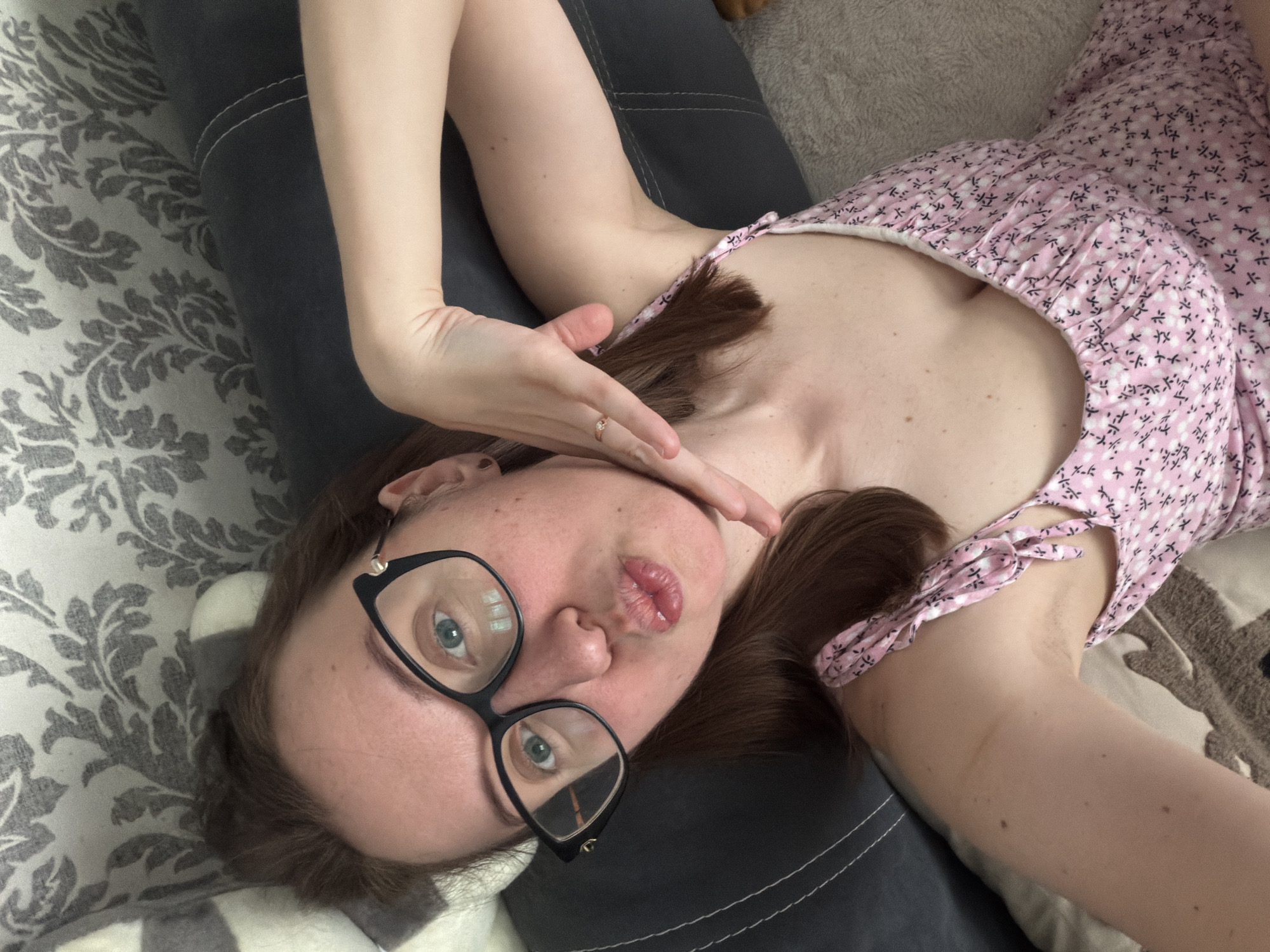 A young woman with brown hair and glasses is lying on her side, looking at the camera with her lips pursed. She is wearing a pink floral dress and a ring on her left hand. The background is a couch with a patterned blanket. The image appears to have been taken in a residential setting, possibly a living room or bedroom. There is no information about the location in Bila Tserkva, Ukraine.