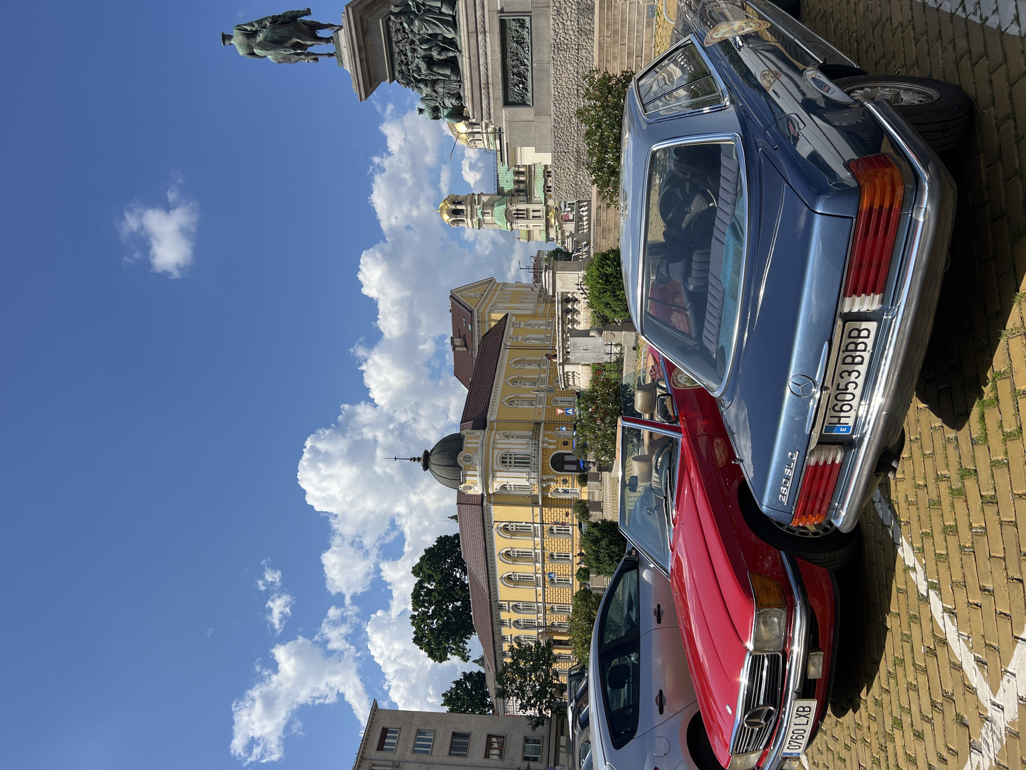 The image captures an outdoor urban scene in Sofia, Bulgaria, on a clear, sunny day under a blue sky with scattered white cumulus clouds. In the foreground, two vintage Mercedes-Benz automobiles are parked on a yellow brick-paved surface with white parking lines; grass grows between the bricks. On the left is a red Mercedes-Benz R107 SL convertible, displaying the Bulgarian license plate "0760 LXB". To its right, a blue Mercedes-Benz C107 280 SLC coupe bears the Bulgarian license plate "H6053 BBB" and has a circular emblem on its side. A silver hatchback is partially visible behind the red Mercedes.
In the midground, to the left, stands a plain, light-colored multi-story building. Center-left, a large, ornate yellow building with a dark dome and numerous arched windows is visible, surrounded by trees and shrubbery. A path or road runs in front of this building. To the right, a multi-tiered stone pedestal supports a bronze equestrian statue, the Monument to the Tsar Liberator (Alexander II of Russia), which is seen from behind. Bas-reliefs depicting multiple figures adorn the lower part of the monument. Stairs lead up to the monument area, and green bushes are interspersed throughout.
In the background, positioned right of center, the St. Alexander Nevsky Cathedral is prominent, characterized by its distinctive golden domes and green-patina roofs, partially obscured by the monument and surrounding foliage. Several indistinct figures are visible near the yellow building and on the steps leading to the monument.