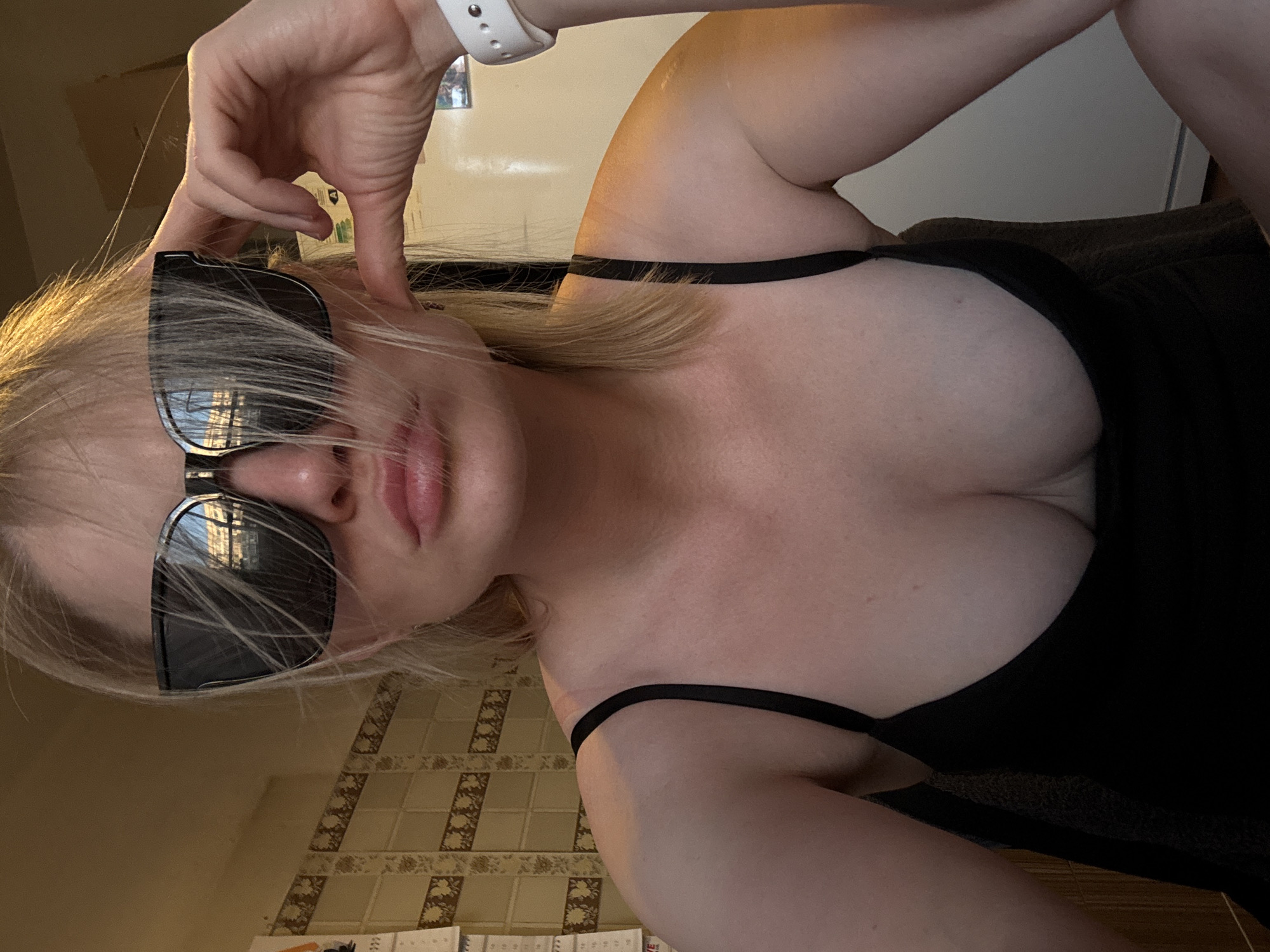 The image features a young adult female, likely taking a selfie indoors. She has blonde hair, which partially covers her face and black sunglasses. Her lips are slightly parted. She is wearing black thin-strapped clothing, possibly a top or a dress, which reveals her décolletage. A white, perforated wristband, resembling a smartwatch or fitness tracker, is visible on her right wrist. Her right hand is raised near her head, forming a gesture with her fingers.
The setting appears to be an interior space, with a wall featuring a pattern of light brown and beige tiles visible in the lower left portion of the frame. Another plain, light-colored wall or door is seen in the background. A dark brown, textured surface, possibly upholstery or carpet, is also visible. Reflections in her sunglasses suggest an outdoor view, possibly buildings or windows, indicating natural light.
The photograph is taken from a low angle and is rotated 90 degrees clockwise. The lighting suggests it is daytime. There is no discernible text visible in the image. While the provided context indicates Saint Petersburg, Russia, there are no specific visual cues within the image itself to confirm this location. The overall mood is casual and relaxed.