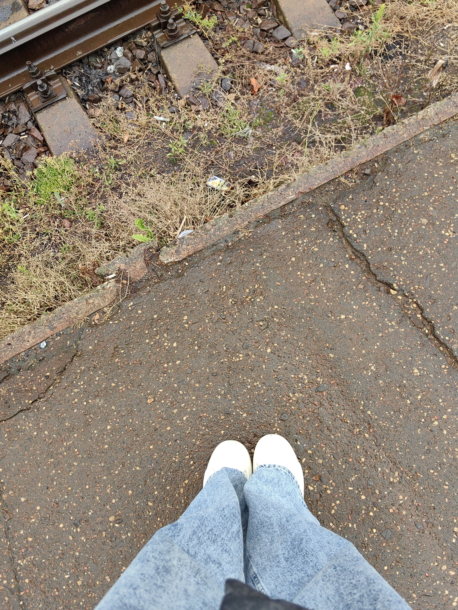 The image shows a person's feet wearing light blue jeans and white shoes standing on a cracked, wet asphalt surface. To the left of the asphalt is a rusty metal border and beyond that, railroad tracks embedded in gravel and dry grass. The scene suggests an outdoor environment, possibly near a railway line in Fanipal, Belarus.