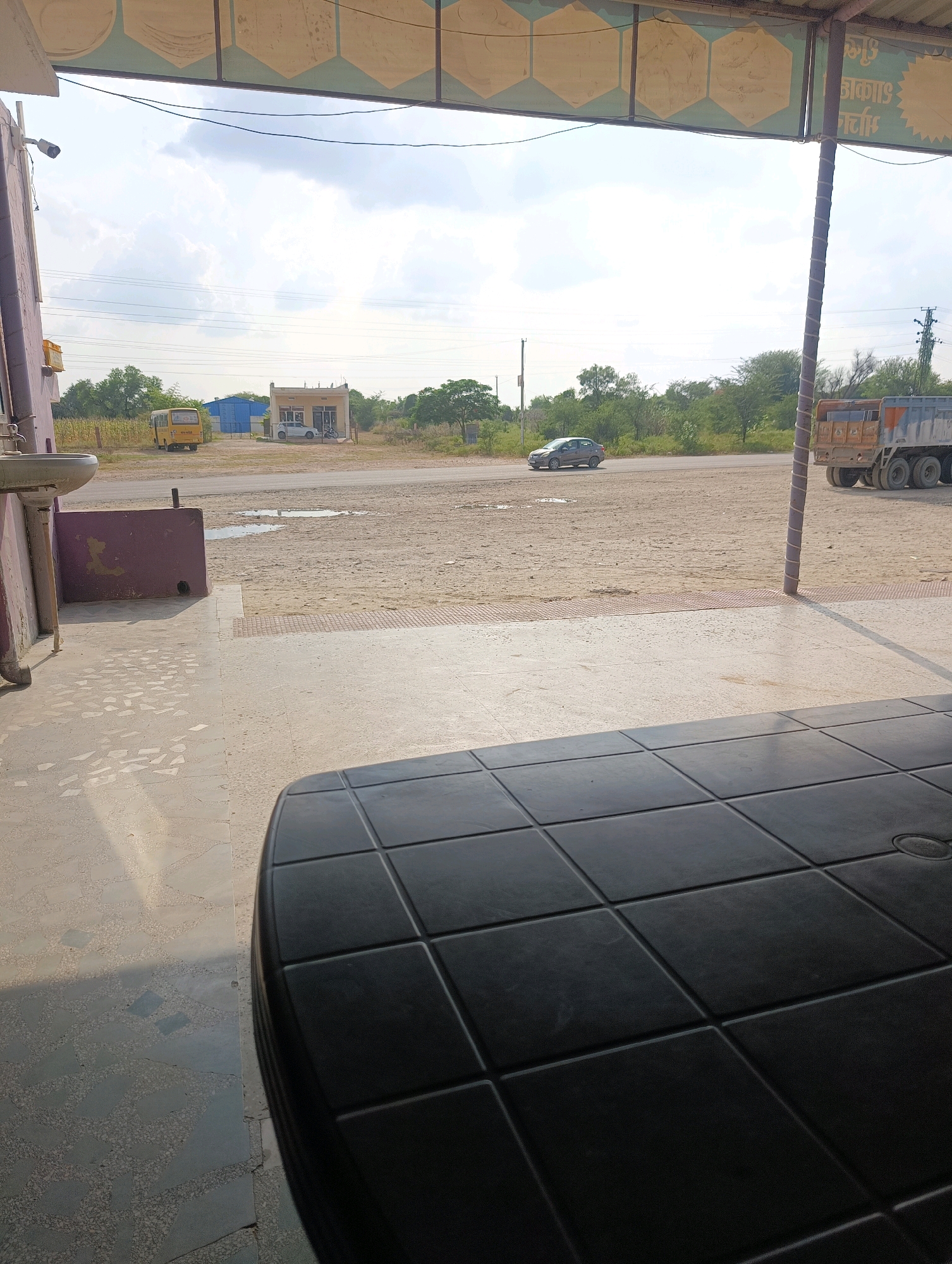 The image captures a scene in Chhapri Bari, India, from a covered outdoor seating area. In the foreground, a black plastic table with a grid pattern is visible, partially obscuring the floor which is tiled with a mosaic of light gray and darker gray geometric shapes. Beyond the seating area, a dusty, unpaved ground extends to a road. On the road, a silver sedan is driving away from the viewer, and further down the road, a large, rusty dump truck is parked. In the background, several buildings are visible: a small, light-colored commercial building with a blue roof behind it, and a yellow school bus parked near the buildings. The sky is cloudy with a bright overcast. The overall impression is that of a roadside establishment, possibly a dhaba or a rest stop, in a rural or semi-urban Indian setting.