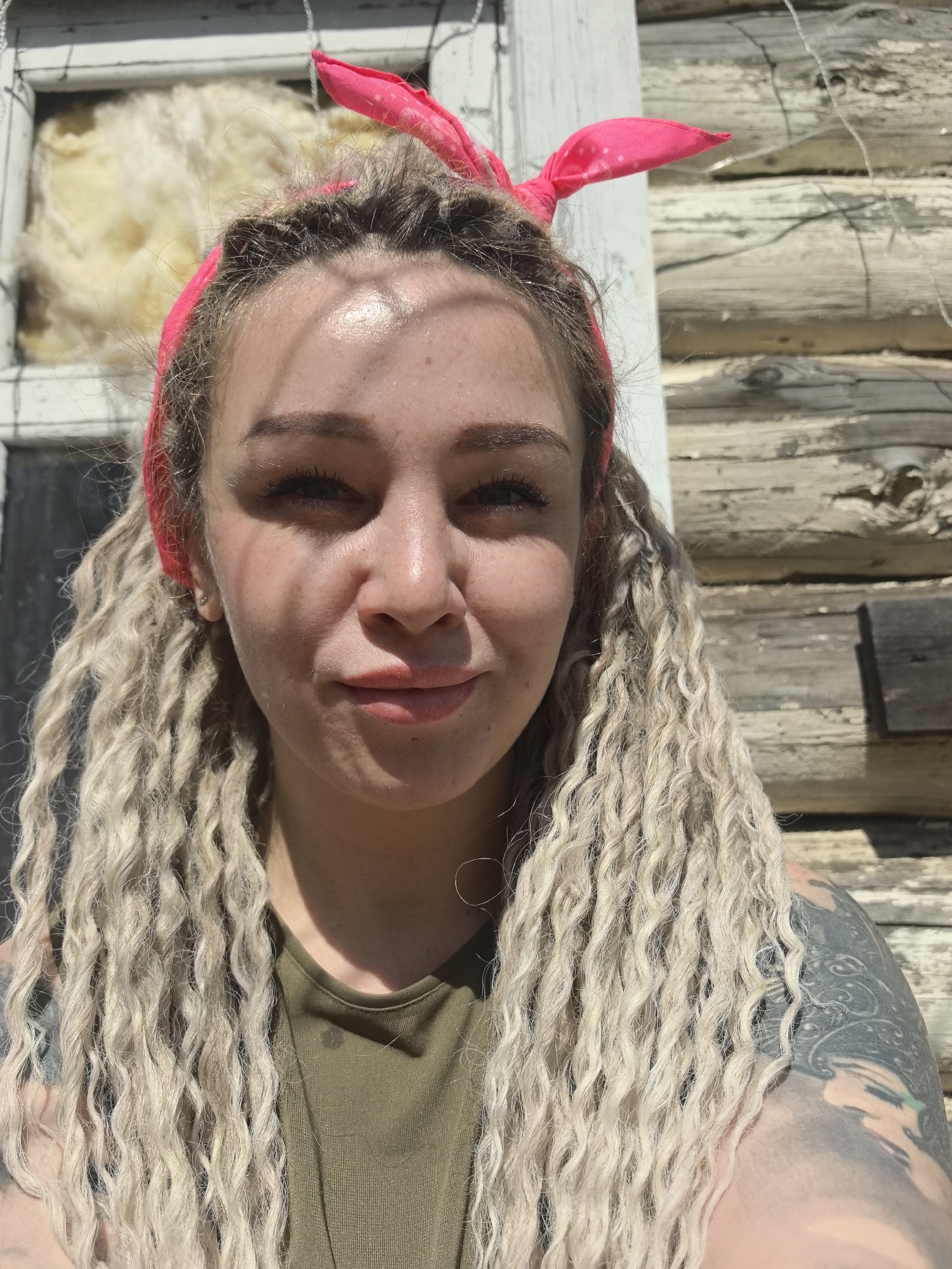 A woman is standing outside in front of a log cabin. She has light skin, curly blonde hair, and a pink bandana. She is wearing a green shirt. She has a tattoo on her arm. The location is not stated, but the image does include the city of Noviy, Russia.