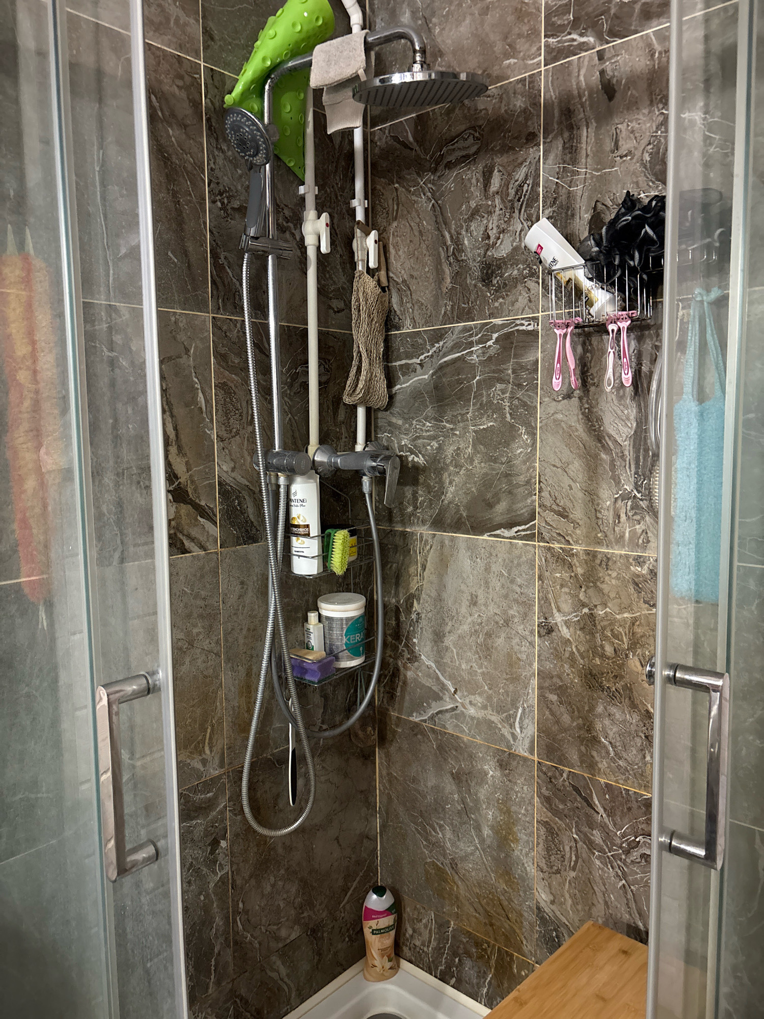 The image shows a shower stall with marble-patterned tiles. A shower head, hand-held shower wand, and various toiletries are visible. Among the items are bottles of shampoo and body wash, razors, and loofahs. A wooden bench is positioned near the shower floor. The shower stall has glass doors with chrome handles. The overall impression is one of a functional and well-stocked shower area.