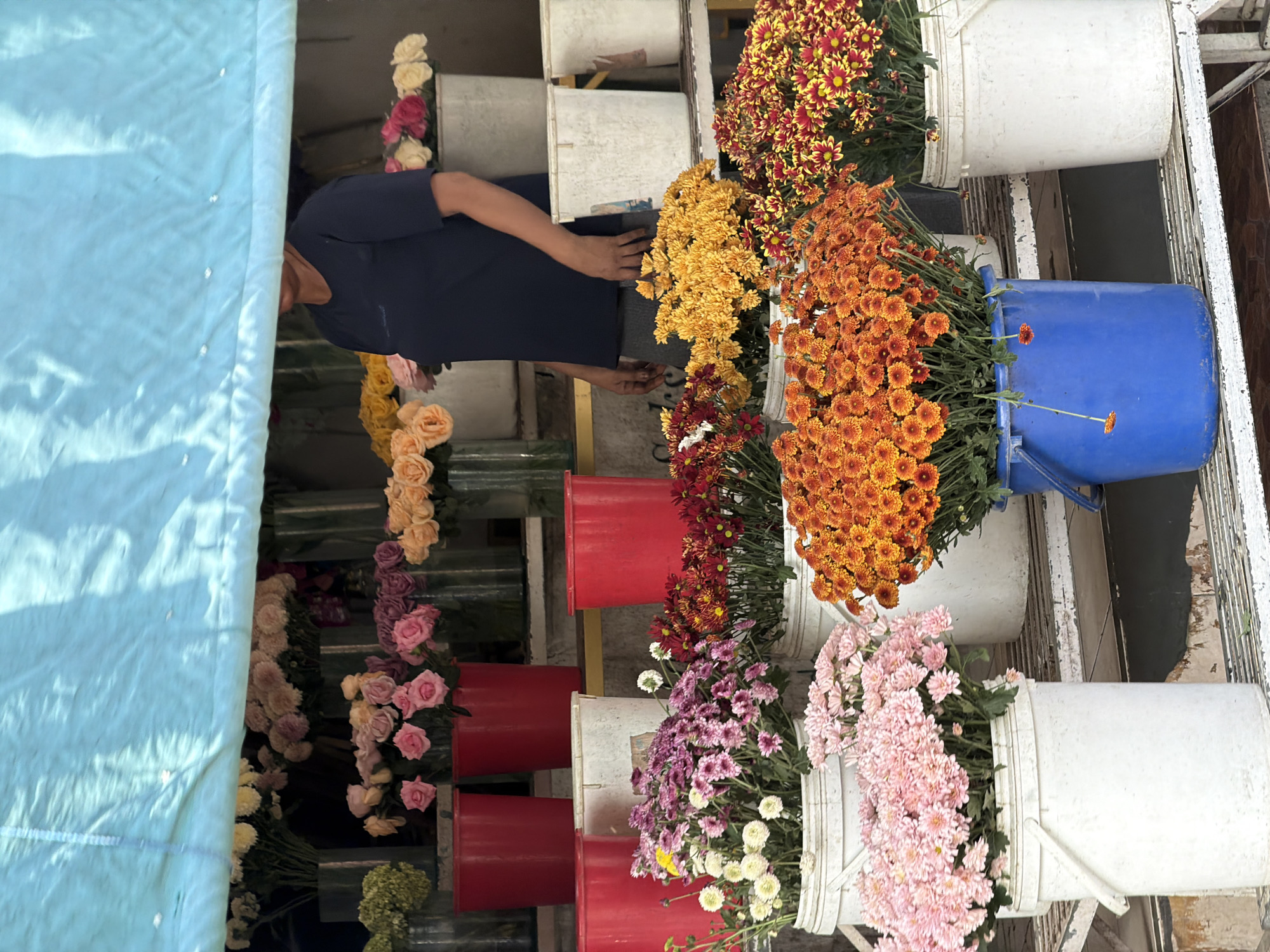 A vendor stands behind a flower stall under a blue tarp, displaying a variety of flowers in buckets. The stall is positioned outdoors, likely on a street in the Ubud District of Indonesia. The vendor, wearing a dark blue t-shirt and gray pants, is partially obscured by the flowers. The flowers include pink, white, yellow, orange, and red chrysanthemums, as well as roses in various shades. Buckets of different sizes and colors, including red, white, and blue, are used to hold the flowers. The stall appears to be set up on a tiered shelving unit. The overall scene depicts a vibrant flower market.