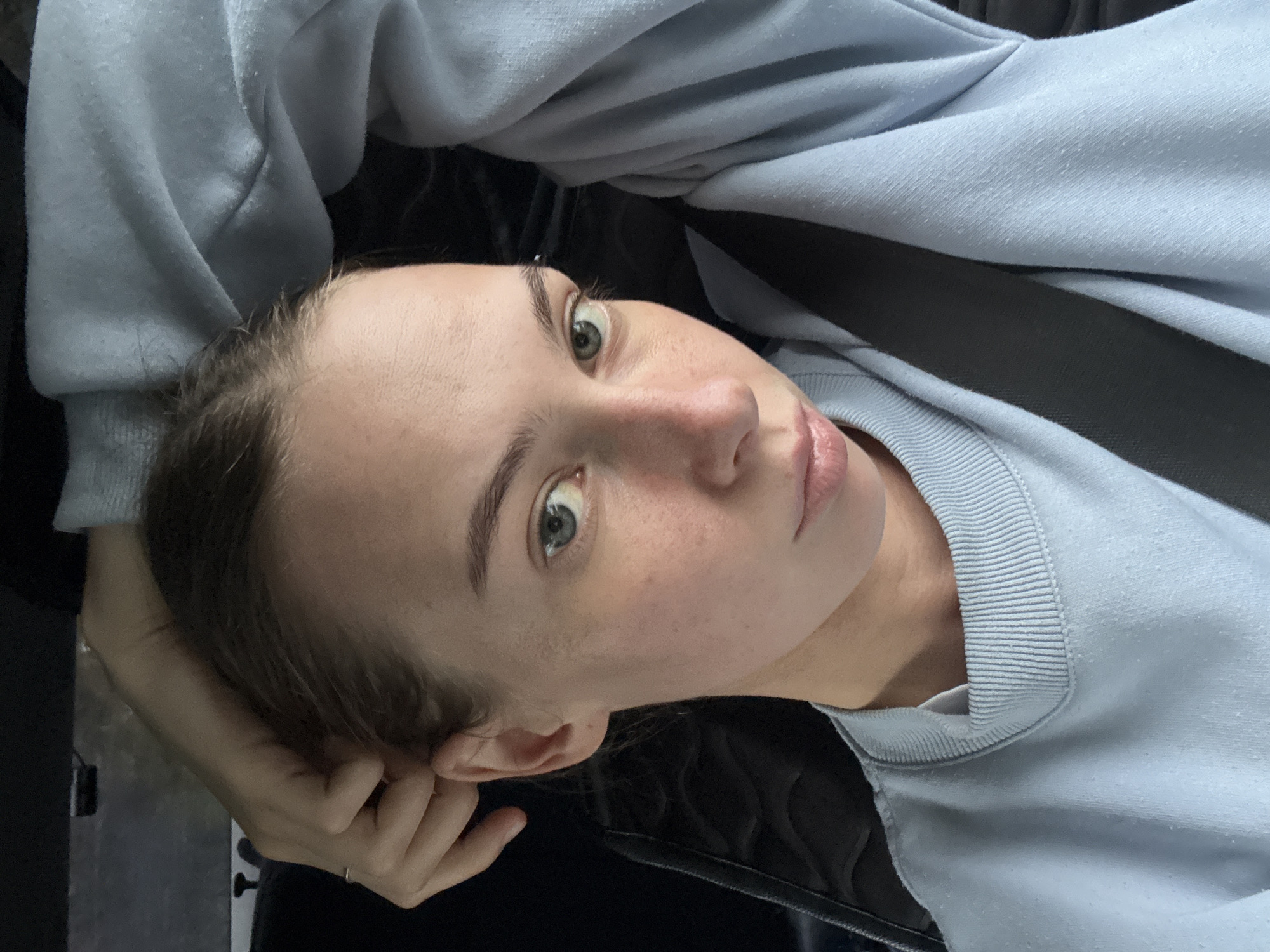 A young woman with light skin and light brown hair, pulled back in a braid, is shown in a close-up, over-the-shoulder shot. She is wearing a light blue crewneck sweatshirt and a black seatbelt. Her left arm is bent, with her hand resting on her head, and her right arm is not visible. Her expression is neutral, with her gaze directed towards the camera. The background is dark and out of focus, with a hint of black upholstery visible on the left. The image appears to have been taken inside a vehicle. The location is the city of Perm, Russia.