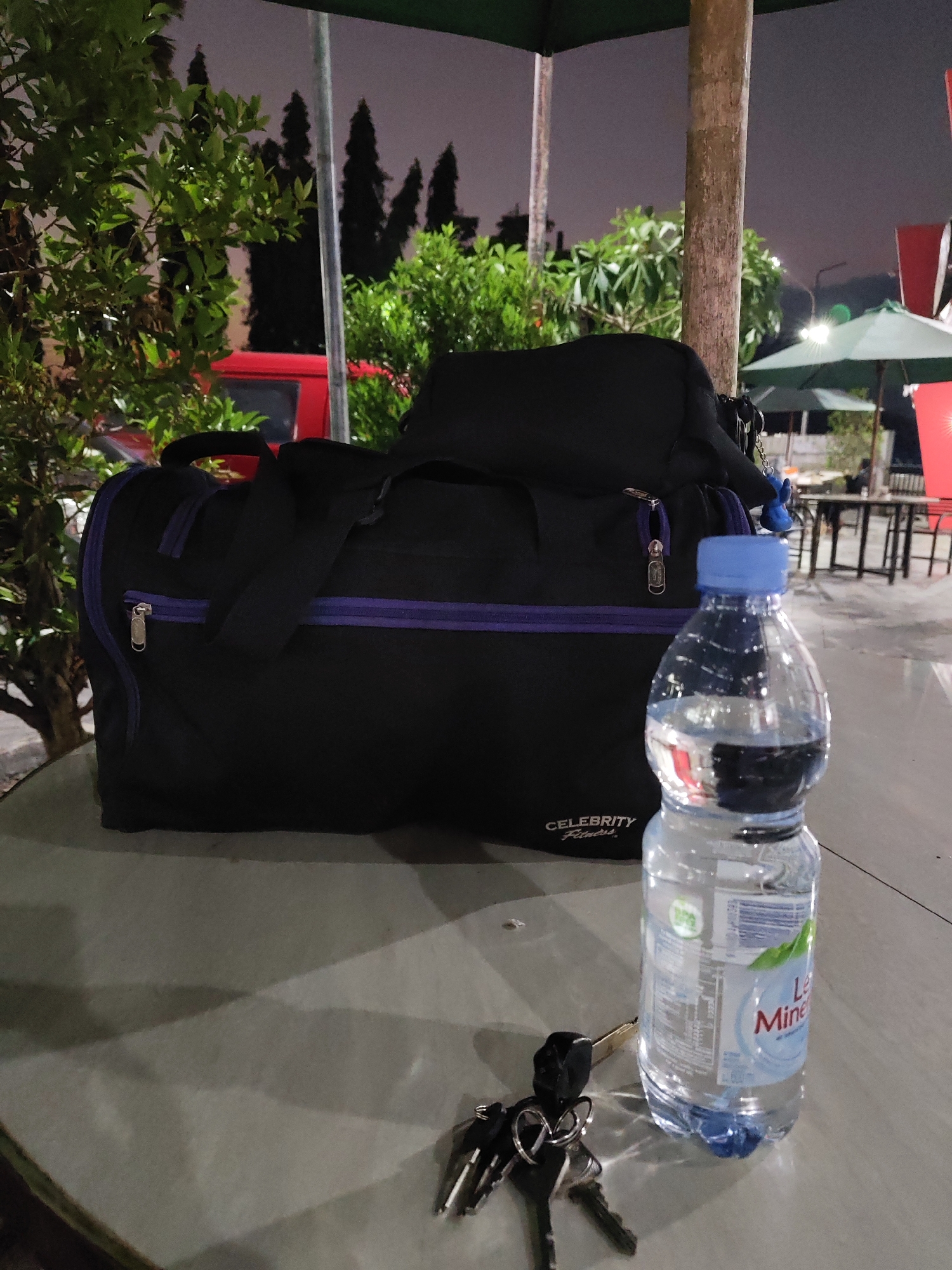 This media file captures a still life scene at night, likely in Geneng, Indonesia. The foreground is dominated by a table on which rests a large, black duffel bag with purple trim. Attached to the bag is a small, blue keychain charm shaped like a bird. Next to the duffel bag is a clear plastic bottle of Le Minerale bottled water, its blue cap prominent. In front of the bottle and bag are a set of keys, including a car key fob.
The background reveals a dimly lit outdoor setting. Lush green trees and foliage frame the left side of the image, with a sliver of a red vehicle visible behind them. In the distance, silhouetted against the dusky sky, are tall, conical trees, possibly pine or cypress. Further back, outdoor seating areas with tables and green umbrellas are visible, suggesting a restaurant or rest stop. The overall impression is one of a brief pause or stop during travel at night. The lighting is artificial, cast by unseen sources, creating shadows on the table. The sky is a dark, muted grey, indicating nighttime. The overall mood is calm and perhaps a little weary, typical of a late-night stop.
Visible text on the duffel bag reads "CELEBRITY Fitness". The water bottle label shows the brand name "Le Minerale".