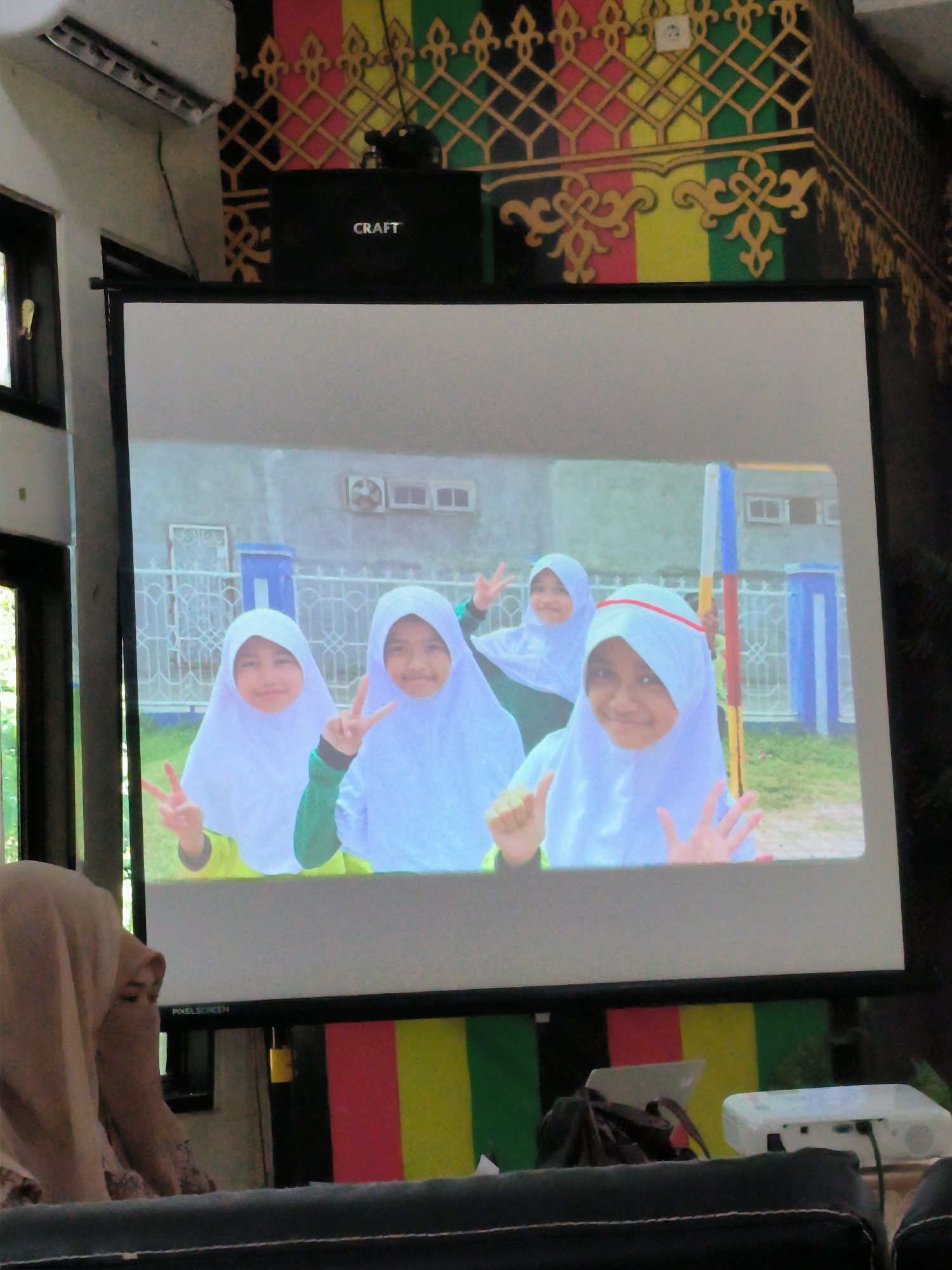 An indoor scene, likely a presentation or meeting room, featuring a large projection screen displaying an image of four young females, located in the city of Unknown, Unknown.
The central focus is a white, rectangular projection screen, framed in black, with "PIXELSCREEN" visible at its lower edge. The screen projects an image of four smiling young females, each wearing a white headscarf (hijab) and colorful long-sleeved shirts (two in yellow, one in green, and one partially obscured). Three of the females are making a peace sign with their fingers, while the fourth shows open palms. The background of the projected image appears to be an outdoor setting with a light-colored building, a fence with blue and white posts, and green grass. An external air conditioning unit is visible on the building in the projected image.
Above the projection screen, a black speaker box labeled "CRAFT" in white letters is mounted. To the left of the speaker, a white wall-mounted air conditioning unit is visible. The wall above the screen is adorned with intricate, colorful geometric and flowing patterns in red, yellow, green, and black. A white electrical outlet is positioned on this decorated wall.
In the immediate foreground, to the bottom left, the top of a person's head and shoulder are visible. This individual is wearing a light beige or brown headscarf and a patterned garment, facing towards the screen. To the bottom right, a white projector unit with visible cables is situated on a surface, likely a table. A dark, upholstered surface, possibly the back of a chair or sofa, occupies the bottom edge of the frame. On the far left, a dark-framed window reveals a glimpse of green foliage outside.