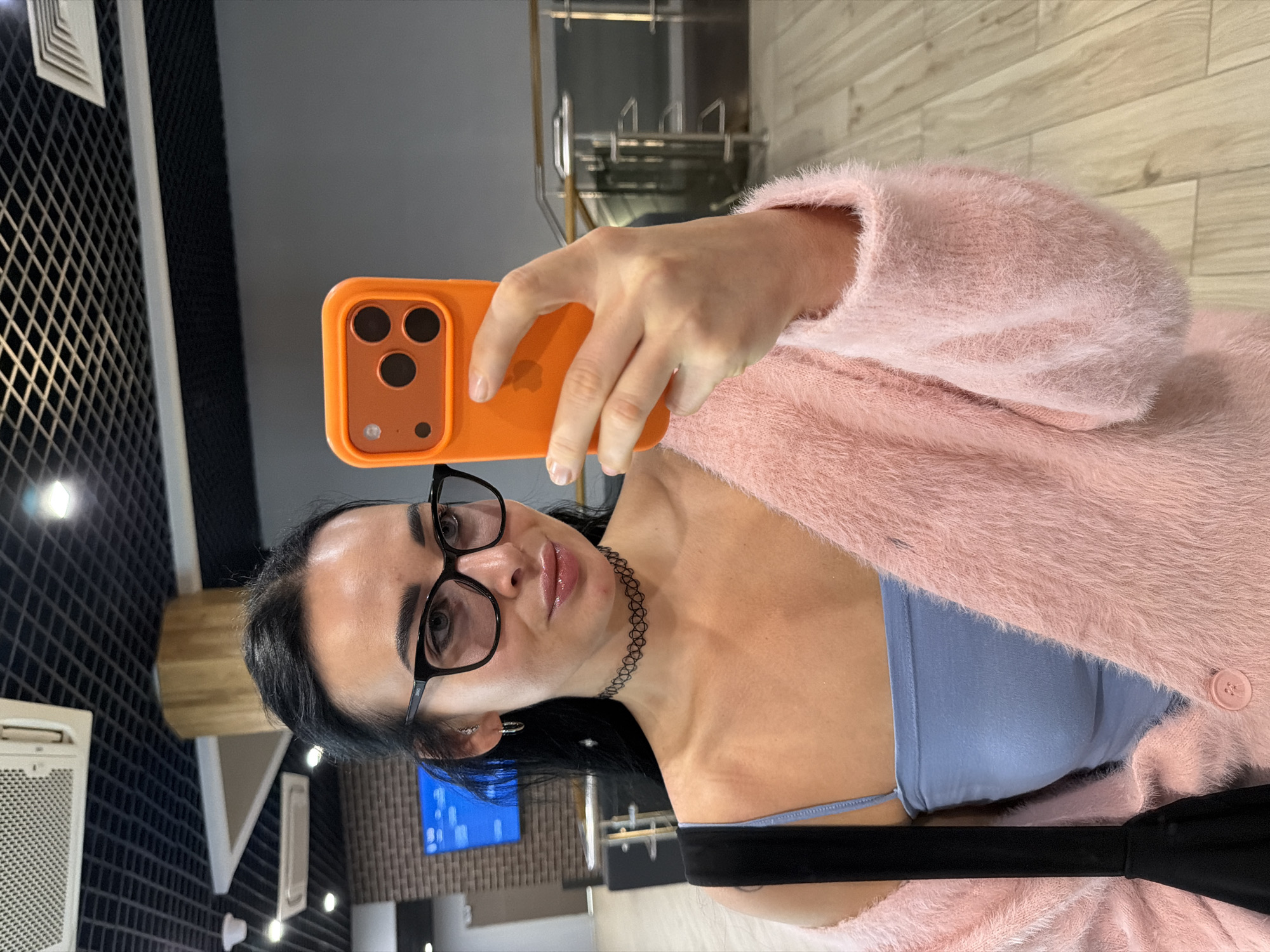 A woman with dark hair and glasses is taking a selfie with an orange phone. She is wearing a blue top and a pink fuzzy cardigan. She has a black choker necklace. The background appears to be an interior space with gray walls and a tiled floor. There is a black wall with a white vent. A screen and chrome fixtures are visible. The location is likely indoors and does not appear to be in Saint Petersburg, Russia.