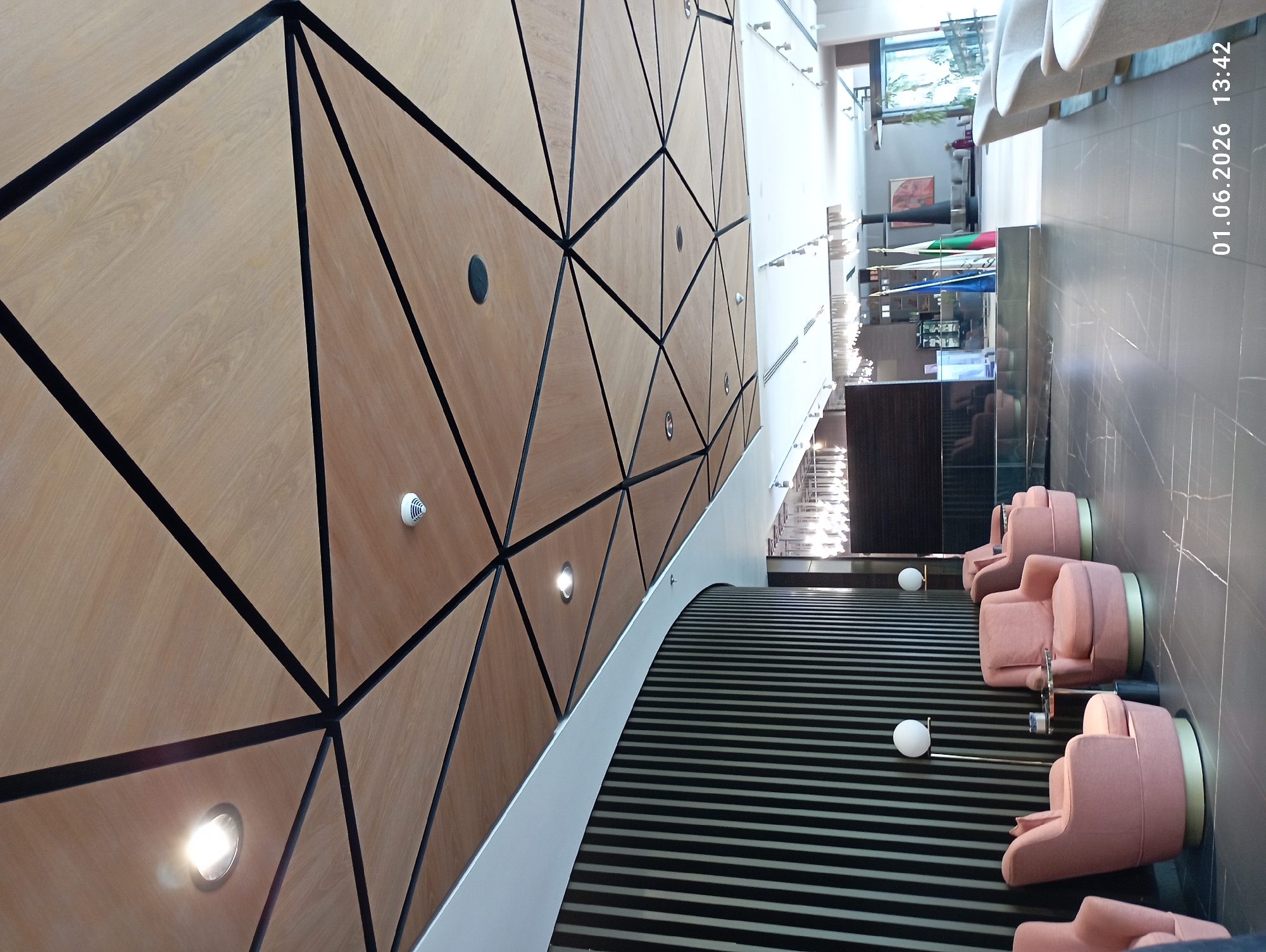 The image depicts an interior space with a geometric wall design on the left. The wall is clad in light-colored wood panels separated by black lines that create triangular and polygonal shapes. Several small, round lights are embedded within the panels. To the right of the wall is a striped carpeted area, and beyond that, a tiled floor with pink upholstered chairs and small tables. In the distance, there is a bar area with bottles and glasses visible. The scene appears to be a modern hotel lobby or lounge. The location is in the city of Unknown, Unknown. There are no people in the image.