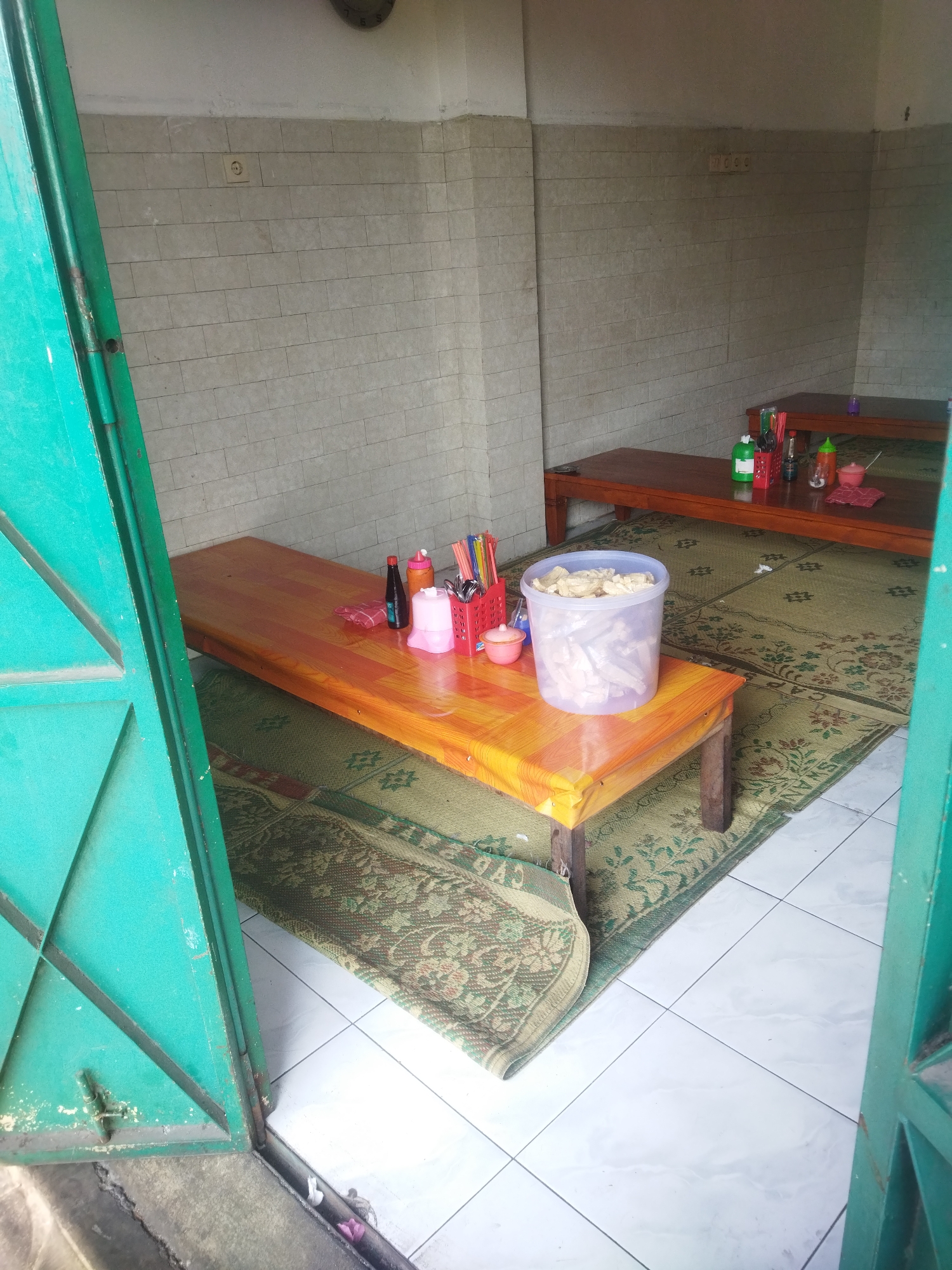 The image depicts a casual dining area, possibly a simple restaurant or food stall, in Wonogiri, Indonesia. The setting is rustic and practical, with tiled walls and floor. Two wooden tables are visible, covered with orange and brown mats, respectively. On the tables, there are various condiments, including bottles of sauce and a plastic container filled with crackers or similar food items. Chopsticks and other utensils are neatly arranged in a red holder. The overall ambiance suggests a place geared towards a quick and informal meal.