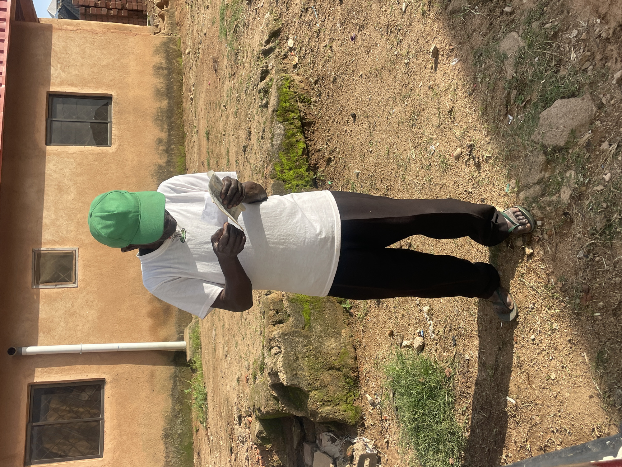 A full-body, eye-level, outdoor, medium shot captures a man standing in front of a light brown, stucco-like building, likely in the city of Barakin Lerawi, Nigeria. The man, wearing a white t-shirt, black pants, and a green baseball cap, is looking down at a small stack of what appears to be money in his hands. He is facing the camera directly. The building has two visible windows on the left, one larger than the other, both with dark panes. A white downspout runs vertically along the side of the building. The ground is covered in dry, dusty earth, with scattered rocks and patches of dry grass. The lighting suggests it is daytime, with clear shadows cast on the ground. To the right of the man, a pile of red bricks is visible, indicating construction or renovation work. The overall scene depicts a moment of financial transaction or review in an unpaved, rural-like setting within a Nigerian urban or semi-urban area.