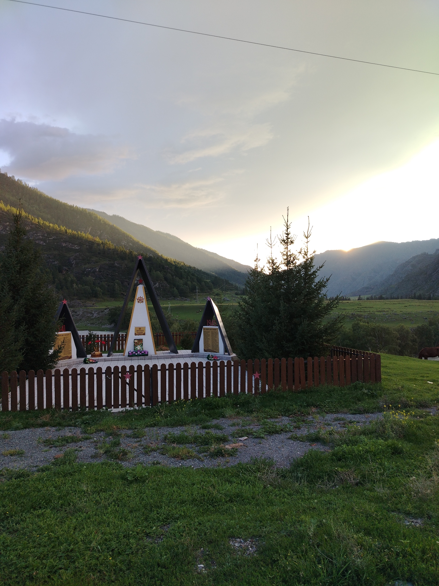 This image captures a memorial with three triangular structures, each topped with a red star, set against a backdrop of lush green hills and a sunset. The memorial is enclosed by a wooden picket fence. The structures appear to be made of concrete and metal, with plaques or inscriptions visible on their surfaces. One of the central structures is adorned with a circular emblem and a floral arrangement. The sky is a gradient of pale blue and orange, with the sun low on the horizon, casting a warm glow. Trees and vegetation are abundant, with a dense evergreen tree to the left and more scattered trees and grassy fields in the distance. A dark brown animal, possibly a cow, is visible in the far right of the image, grazing in the field. The foreground shows a path with gravel and patches of grass. The overall impression is one of solemnity and natural beauty, set in a rural or mountainous landscape. The location in Iodro, Russia, suggests a historical or commemorative site, possibly related to World War II, given the presence of red stars.