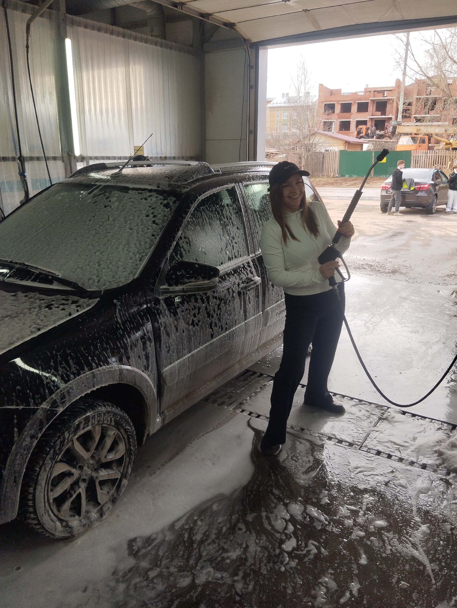 The image shows a woman standing in a car wash facility in Votkinsk, Russia. She is holding a pressure washer and smiling. A dark-colored car covered in foam is to her left. Outside the car wash, two people are standing near a car. A building under construction is visible in the background. The ground is wet and covered in foam. The car wash is an open structure with metal sheeting on the left side. The woman is wearing a white long-sleeved shirt, dark pants, and a baseball cap.