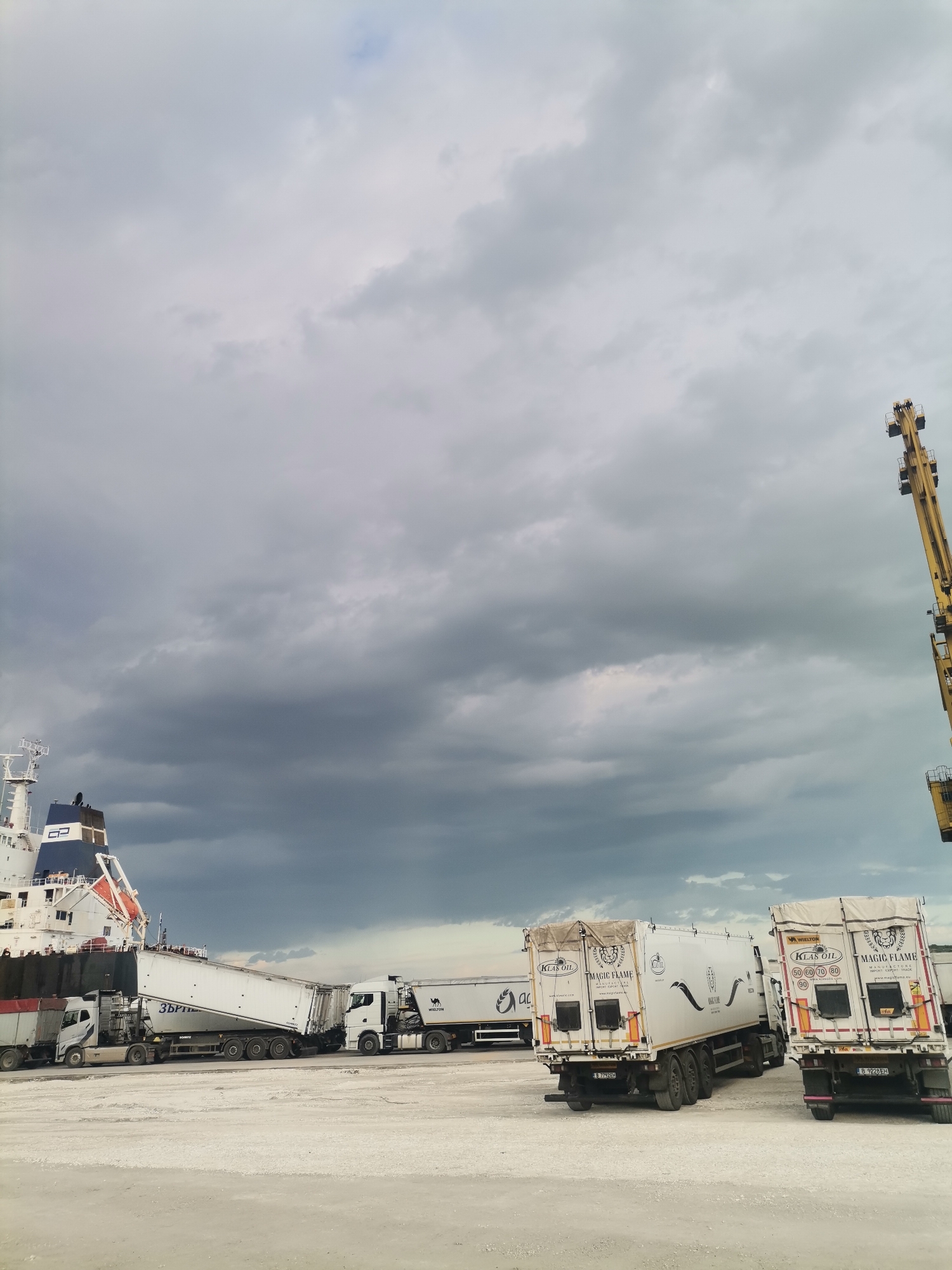The image shows a scene in Ezerovo, Bulgaria. Several large commercial trucks are parked on a light-colored, potentially gravel or concrete surface. The trucks appear to be loading or unloading cargo from a large ship that is partially visible on the left side of the frame. A tall yellow crane is visible in the background, on the right. The sky is overcast with dark, heavy clouds, suggesting an impending storm or recent rainfall. The trucks in the foreground have "KLAS OIL" and "MAGIC FLAME" printed on their rear doors, along with license plates starting with "B." One of the trucks next to the ship has Cyrillic script on its trailer that includes the word "ЗЪР." Another trailer has the word "WIELTON" on its side.