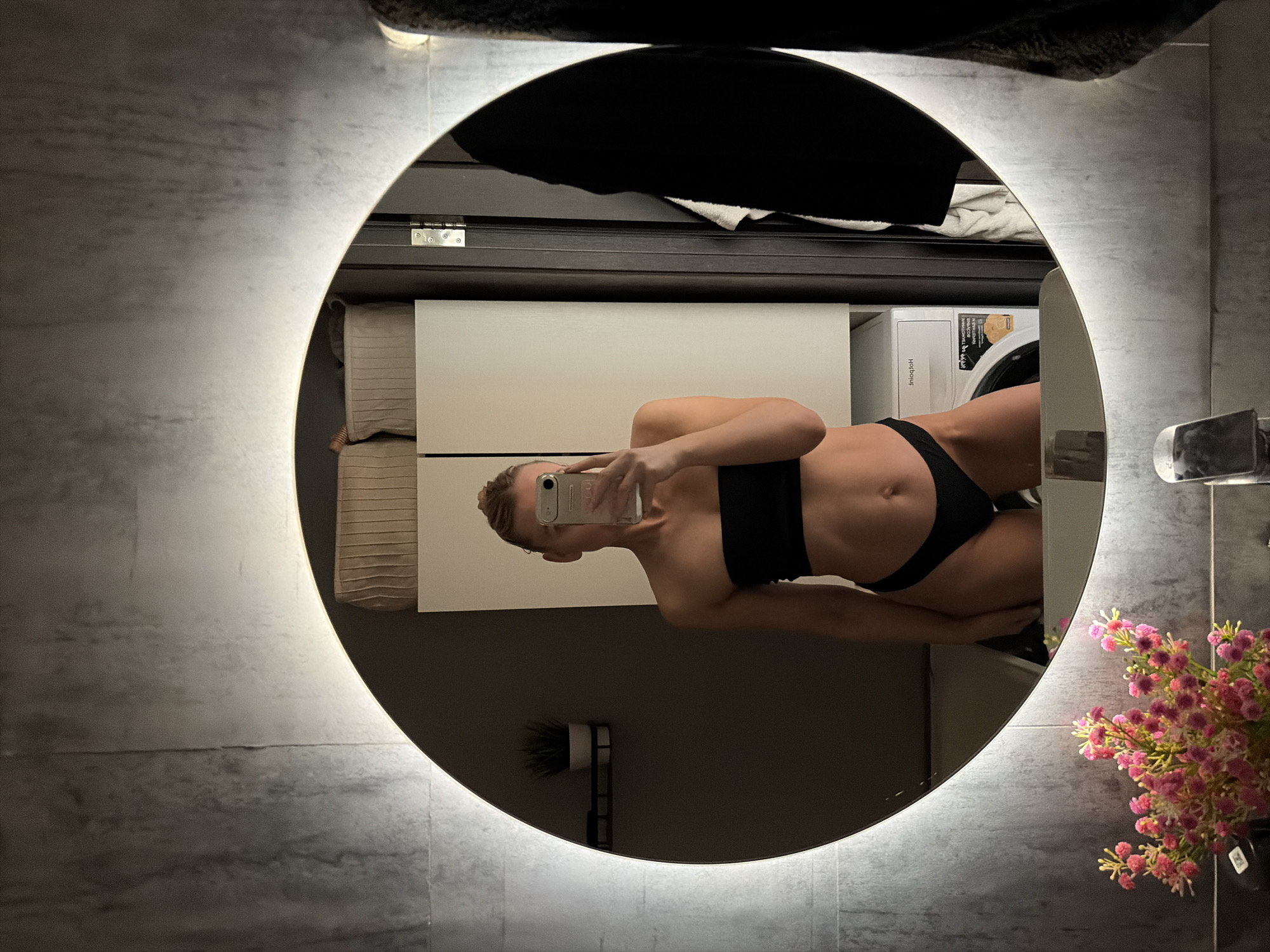 The image is a selfie taken in a bathroom, reflected in a round, LED-backlit mirror. A woman is visible in the mirror's reflection, wearing a black sports bra and black bikini bottoms. She is holding a smartphone up to take the picture. The background shows bathroom elements, including a washing machine, drawers, and items on top of the drawer like folded towels. There is a vase with pink flowers on the right side of the mirror and a small wall mounted decoration on the bottom left side of the mirror. The bathroom walls are tiled in a grey, marble-like pattern. The location is Chaikovsky, Russia.