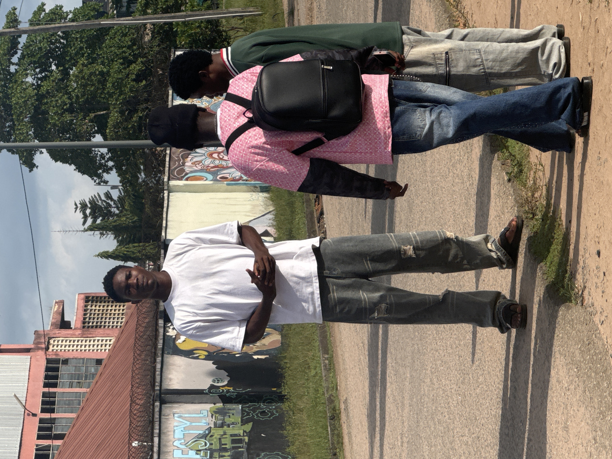 A group of three young men are standing on the side of a road in Ihiagwa, Nigeria. The man in the foreground is wearing a white T-shirt and distressed gray jeans and dark brown sandals. The man behind him wears a pink patterned shirt, blue jeans, a black backpack, and a black beanie. A third man wears a green jacket, gray pants, and black sandals. The setting is an outdoor urban environment with buildings and vegetation nearby. A painted graffiti mural is visible on a wall. The sky is overcast.