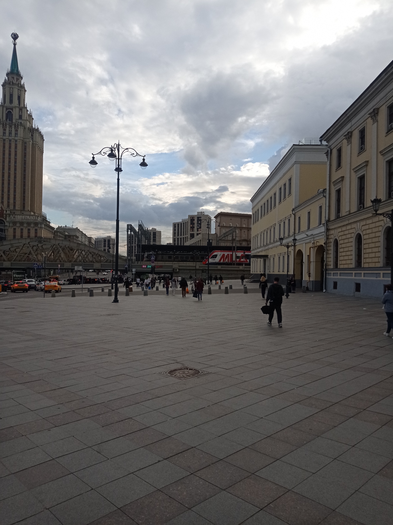 This image depicts an outdoor urban scene in Moscow, Russia, likely near Leningradsky railway station or Komsomolskaya Square, given the presence of the iconic Stalinist skyscraper of the Leningradskaya Hotel on the left and railway tracks in the background. The foreground is dominated by a wide, paved pedestrian area with large, rectangular grey tiles. A street lamp with multiple globes stands prominently in the mid-ground, its silhouette stark against the cloudy sky.
Several people are visible in the scene, mostly walking across the pedestrian area. One individual in the mid-right foreground is walking away from the camera, carrying a dark bag and an umbrella. Other pedestrians are scattered throughout the plaza and near the traffic.
The background features a mix of architectural styles. To the left, the imposing Leningradskaya Hotel, a distinctive Stalinist skyscraper, rises towards the sky. In the center and behind the pedestrian area, a complex network of railway tracks, bridges, and modern buildings is visible. A red and white train with "РЖД" (Russian Railways) branding is present on the tracks. To the right, a long, pale yellow building with classical architectural elements runs along the edge of the square. Traffic, including several yellow taxis, is moving on a road that runs parallel to the buildings and pedestrian area. The sky is overcast with dramatic, grey clouds, suggesting a cooler, possibly temperate day. The overall impression is of a busy, central urban location.