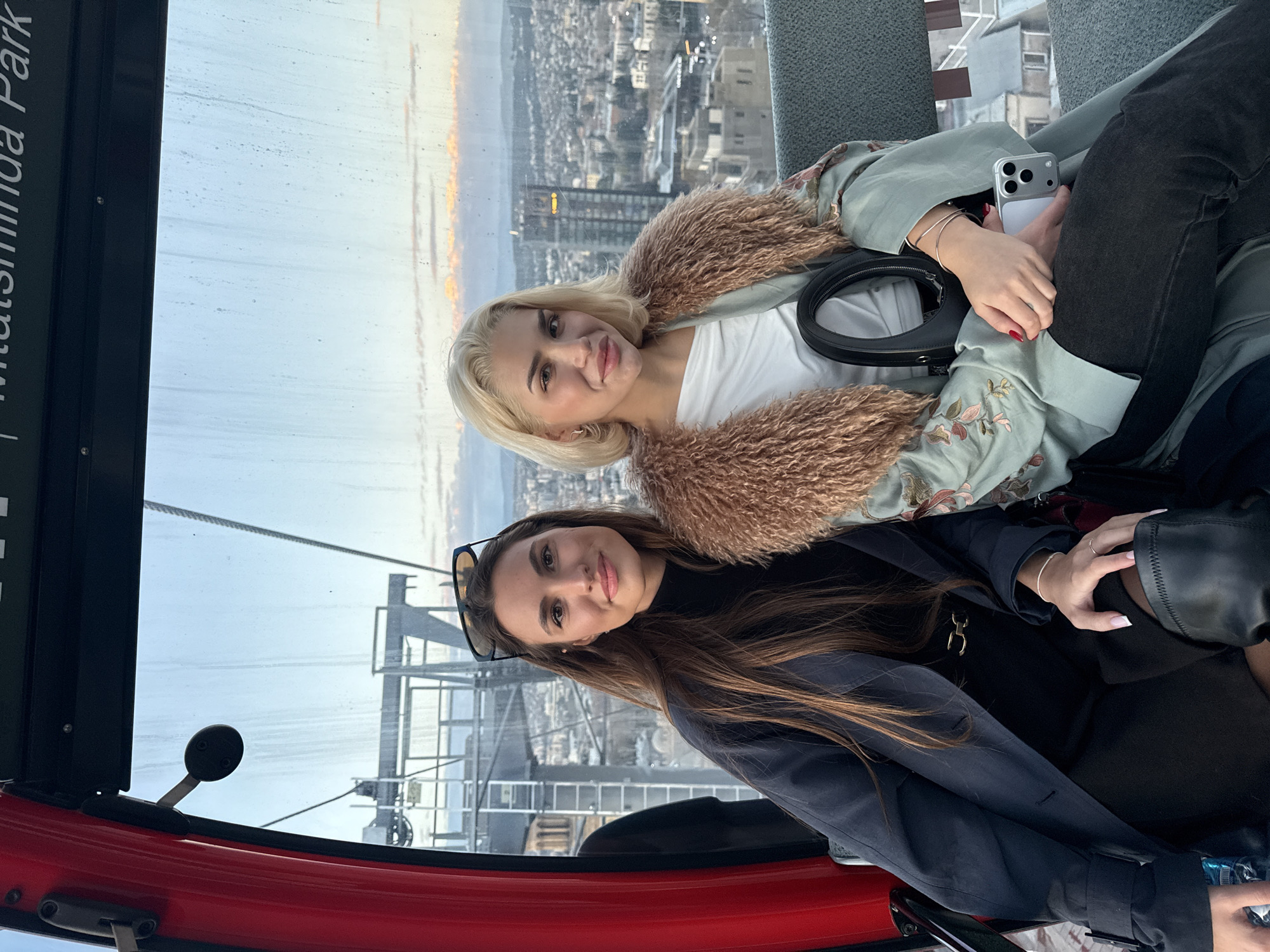 Two women sit inside a gondola car, a cable car with red trim, that is ascending high above Tbilisi, Georgia. The woman nearest the camera has brown hair, is wearing sunglasses, a black top, and a navy jacket. The second woman is blonde, wearing a white top, light green jacket with floral embroidery, and a fur collar. She is holding a silver smartphone. The background reveals a cityscape, a sunset with clouds, and aerial views of buildings and infrastructure. Part of the gondola’s glass panel displays the name "Mtatsminda Park" in white letters.