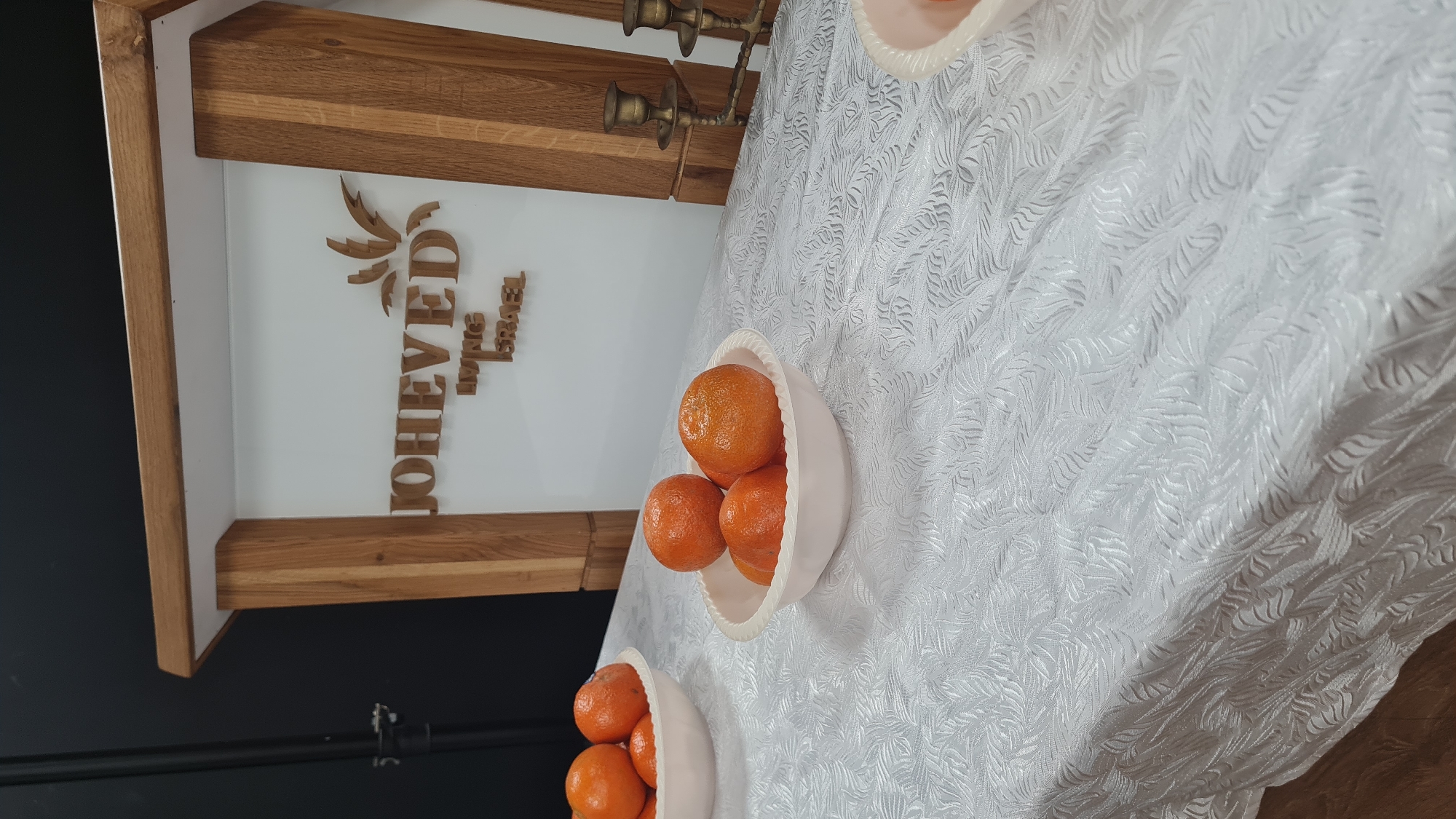 The image shows a table setting in what appears to be a building in Nahariyya, Israel. A white tablecloth with a patterned texture covers the table. On the table, there are bowls filled with oranges. A brass candlestick holder is visible near the table's edge. In the background, there's a sign that says "JOHEVED Living in ISRAEL" mounted on a wall. The overall setting suggests a gathering or event.