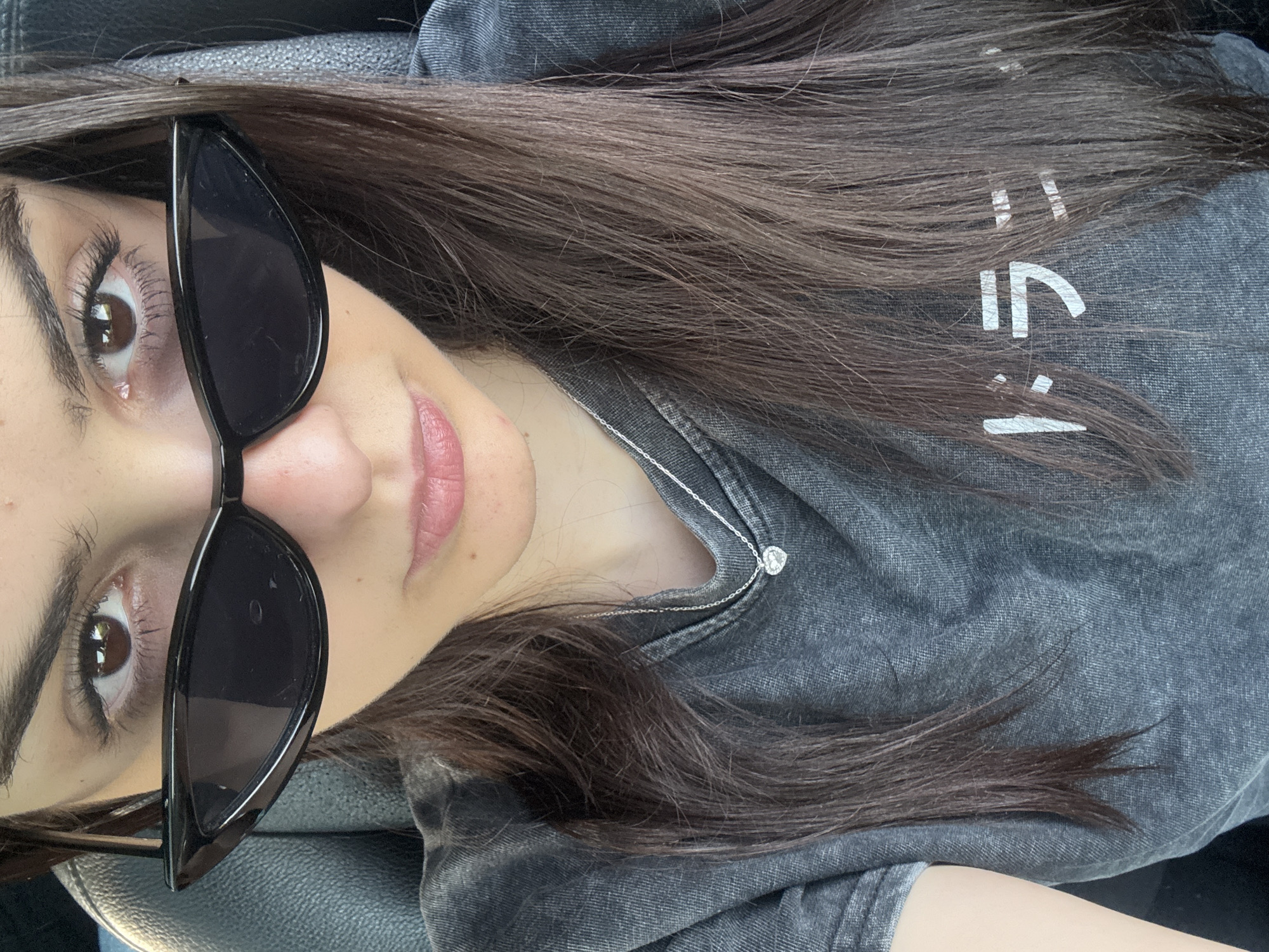 A woman with brown hair and brown eyes is wearing black sunglasses. She is also wearing a gray t-shirt with white lettering and a silver necklace with a round pendant. The image appears to be taken from a slightly elevated, tilted perspective, focusing on her face and upper body. The location is not discernible from the image.
