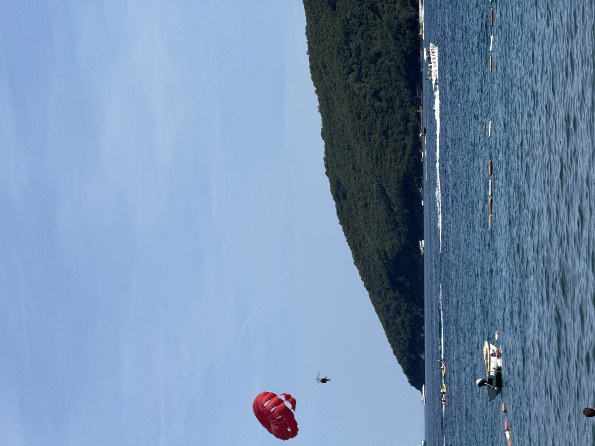 This image depicts a sunny coastal scene in Budva, Montenegro. The sky is a clear, light blue with minimal atmospheric haze. A large, dark green, heavily forested hill or island forms the background, sloping from left to right, meeting the horizon line. The majority of the frame is occupied by the deep blue sea, exhibiting gentle ripples across its surface.
In the upper left quadrant, a bright red parasail is fully inflated and airborne, suspending a single individual in a harness below it. Several boats are active on the water: a white speed boat, marked with partial text 'K-3 P_KYO TD' on its side, moves from left to right in the mid-ground, generating a distinct white wake behind it and carrying a small group of people. Other smaller watercraft, including kayaks, are visible further in the mid-ground. In the foreground, slightly left of center, a white small dinghy equipped with an outboard motor floats stationary. A horizontal line of cylindrical buoys, alternating between orange and white, stretches across the mid-foreground of the water. The top of a person's head is partially visible in the extreme bottom left corner of the frame.