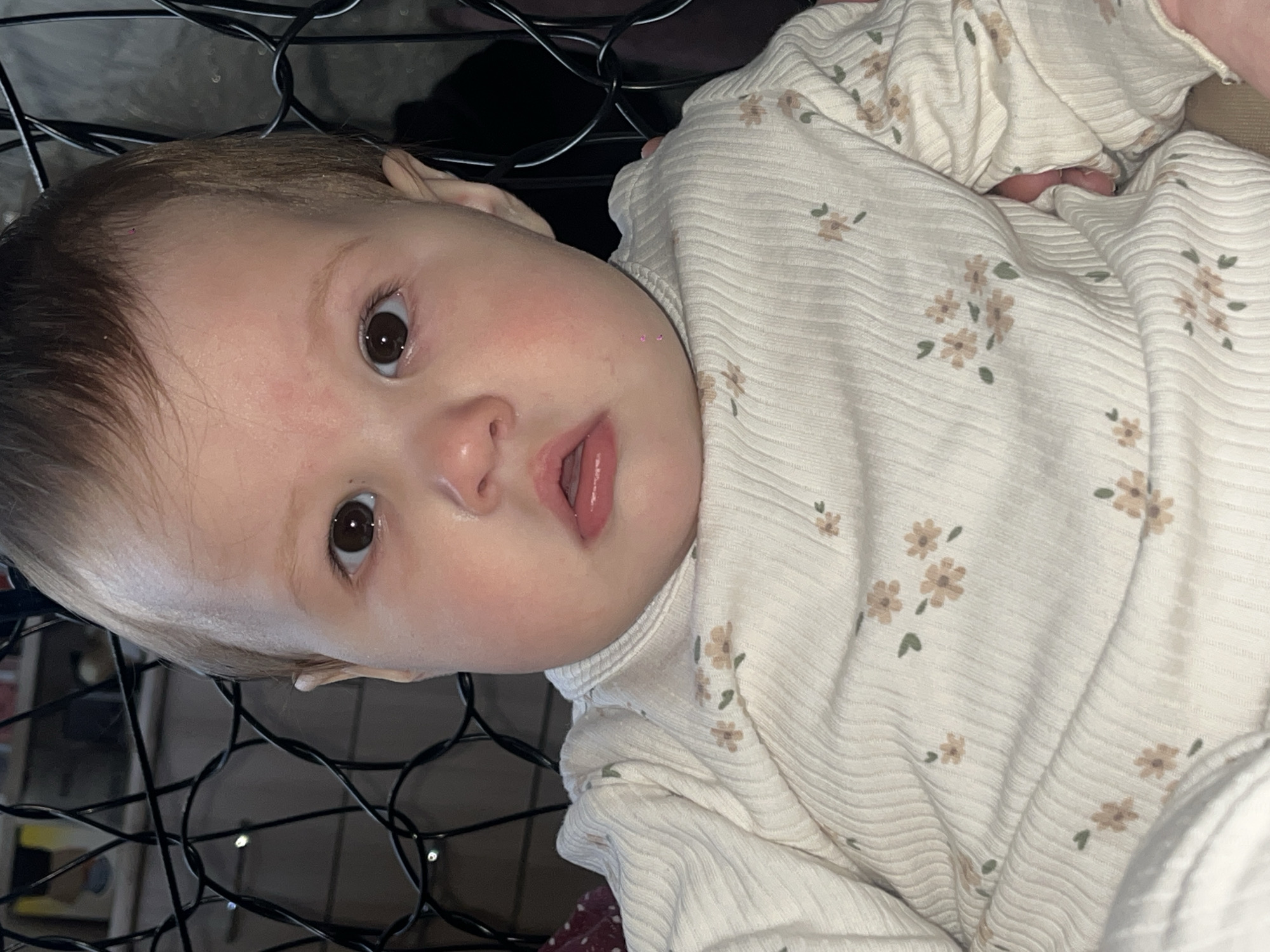A close-up image shows a baby, likely an infant or young toddler, looking directly at the camera. The baby has fair skin, large dark brown eyes, and light brown hair. Small red patches are visible on the forehead and left cheek. The mouth is slightly open, revealing the tongue.
The baby is wearing a cream or off-white ribbed garment, possibly a onesie or shirt, patterned with small, light brown flowers and green leaves. The baby appears to be lying down, with the head tilted.
The background behind the baby's head and to the left is dark and features a prominent black grid-like structure, possibly a crib railing or a decorative element, with black wires intertwined through it. The lighting suggests an indoor setting, likely in a room with dim ambient light, with the baby's face well-lit, possibly by a flash or direct light source.
The baby's expression is alert and curious. No specific activity beyond looking at the camera is depicted. There is no visible text in the image. The location context for this media is Novopokrovskaya, Russia.