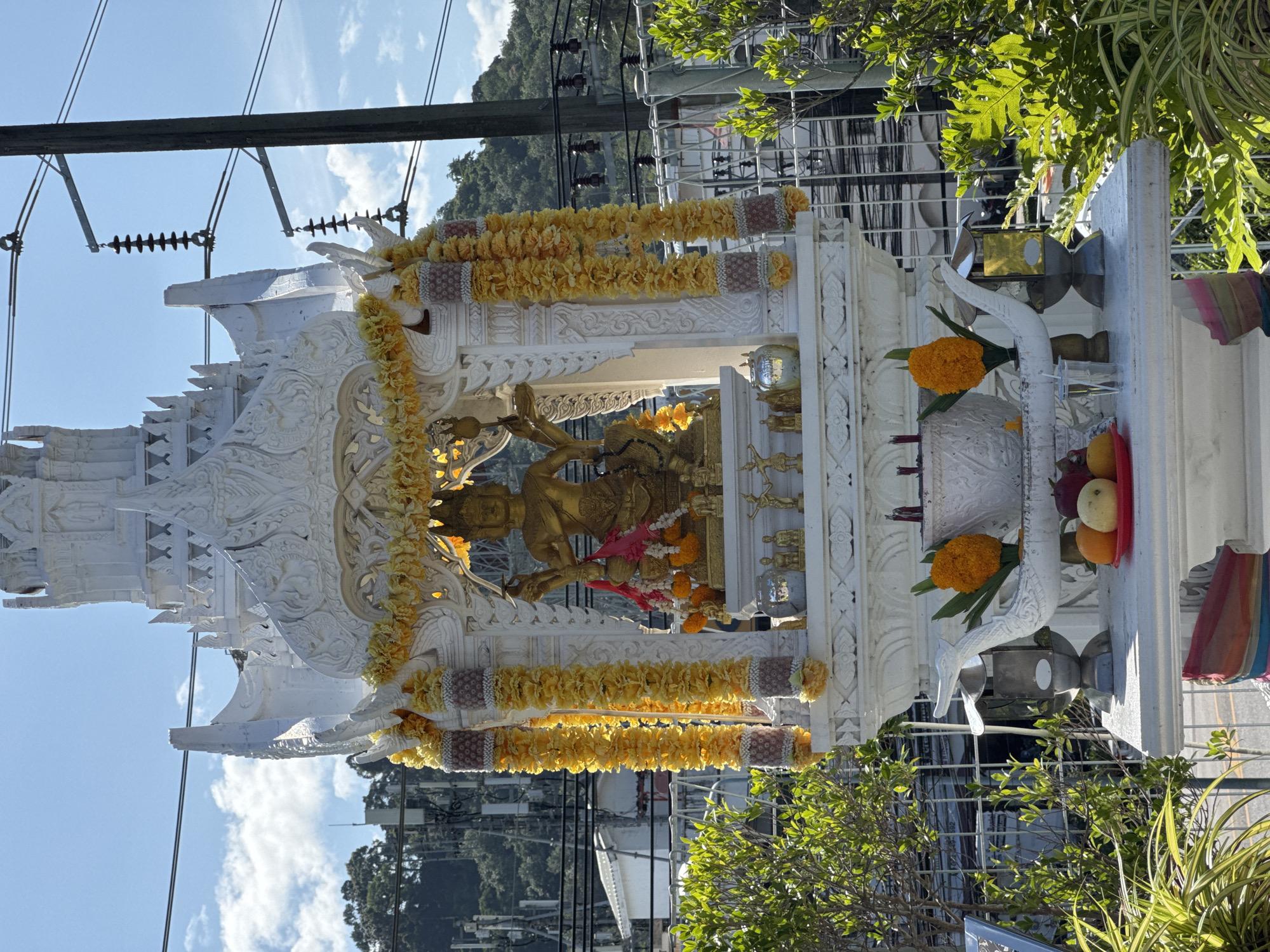The image shows an ornate white shrine, likely a spirit house, with gold decorative elements and yellow flower garlands adorning its exterior. At the center of the shrine, a gold statue of a deity with multiple arms is prominently displayed. Offerings of fruits such as oranges and possibly apples are placed on a red plate at the base of the shrine. Incense sticks are visible in a white bowl. The shrine is situated outdoors, with lush green foliage surrounding it. In the background, power lines and utility poles are visible, indicating an urban or semi-urban setting. The sky is blue with scattered clouds. The overall scene suggests a religious or spiritual site within the city of Wichit, Thailand, as indicated.