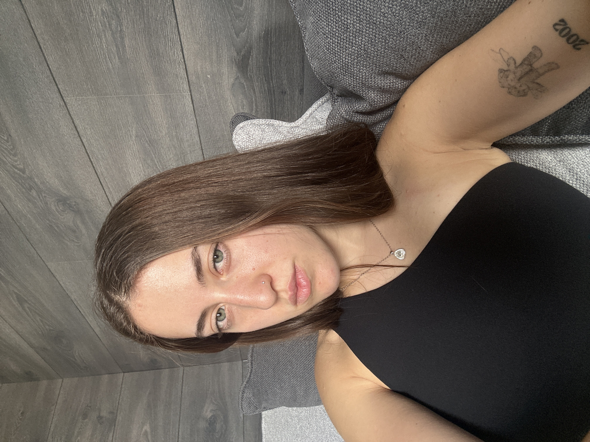A young woman lies on a gray upholstered couch, her head oriented to the left side of the frame. She is wearing a black sleeveless top and a silver necklace with a heart-shaped pendant. Her hair is brown and shoulder-length, and her eyes are green. She has a nose piercing. On her upper left arm, there is a tattoo that appears to include the number "2002". The couch is positioned on a gray wood-look floor. The location cannot be determined from the image.