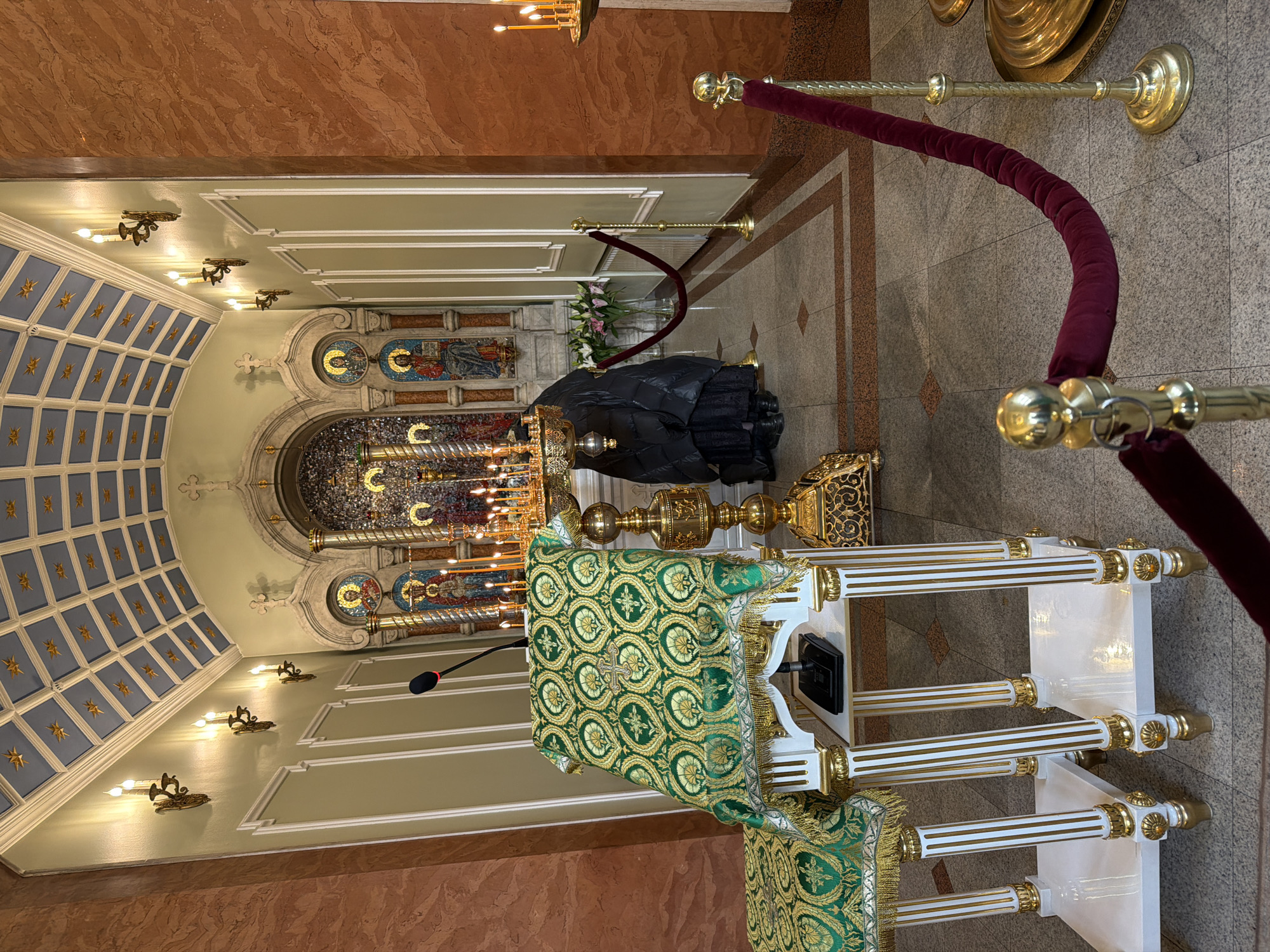 The image depicts an interior view of a church, specifically the burial place of Saint Xenia of Saint Petersburg at the Smolensky Cemetery in Saint Petersburg, Russia. The scene is ornately decorated, characteristic of Orthodox Christian churches. A dark object, likely a coffin covered with a black shroud, is visible in the center of the image, flanked by golden ornate candle stands with lit candles. A green and gold embroidered cloth covers a lectern, placed in front of the coffin. A microphone extends from the lectern, suggesting a service or presentation is being conducted. The walls are pale green, accented with white trim, while the upper walls and ceiling are a darker marble-like color. The ceiling features an arched section with mosaics and Christian symbols, with smaller lights mounted on the wall. A velvet rope barrier is positioned to control the flow of visitors. The floor is light colored tile with a geometric pattern.