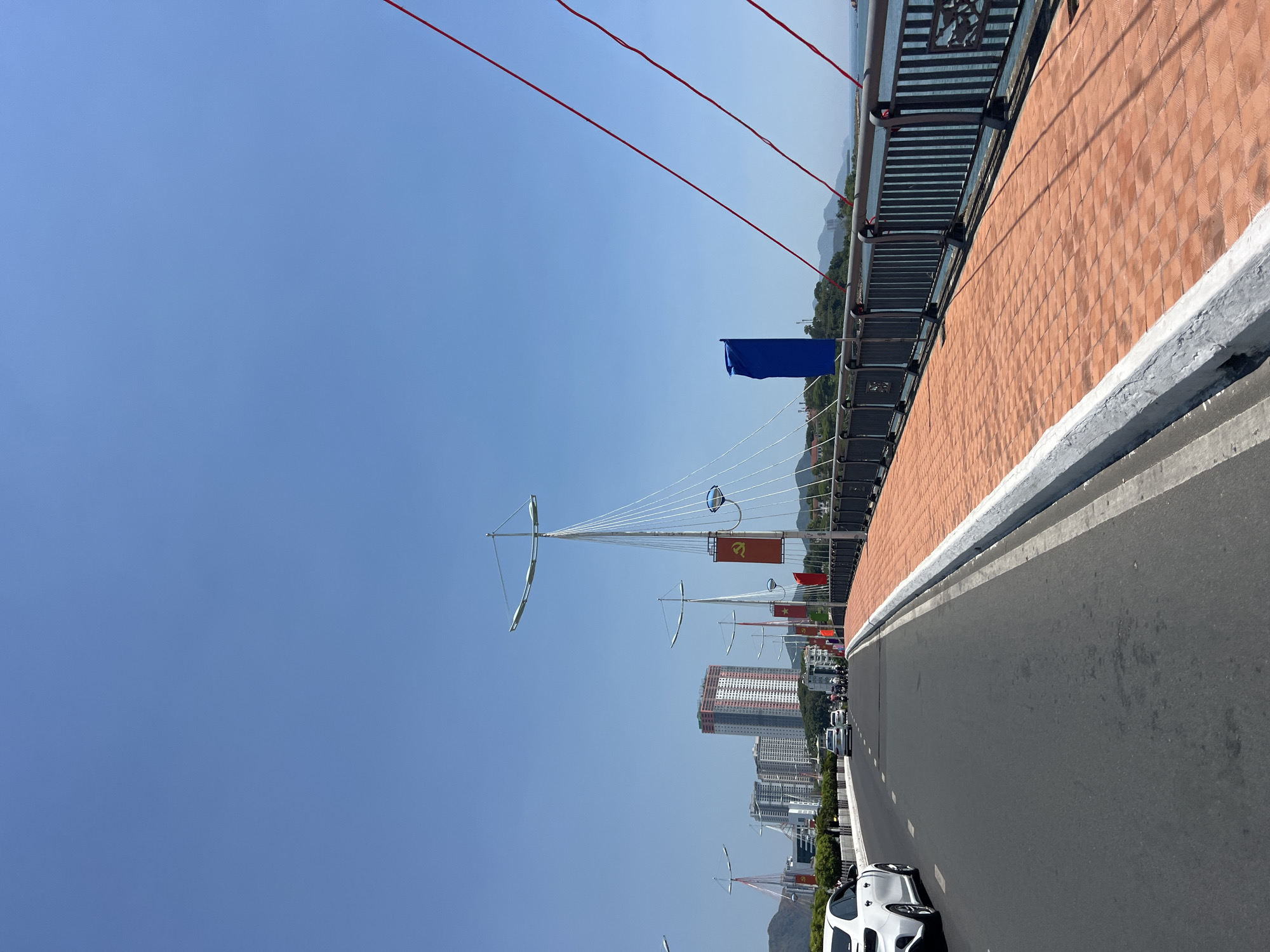 The image depicts a view from a bridge in Nha Trang, Vietnam, looking toward the city skyline. The bridge has a brick sidewall and a railing. Several decorative light poles resembling masts are visible, each adorned with a Communist flag and a blue flag. Cars and other vehicles are traveling on the road along the bridge, which seems to be heading towards a cluster of tall buildings. The sky is clear and blue.