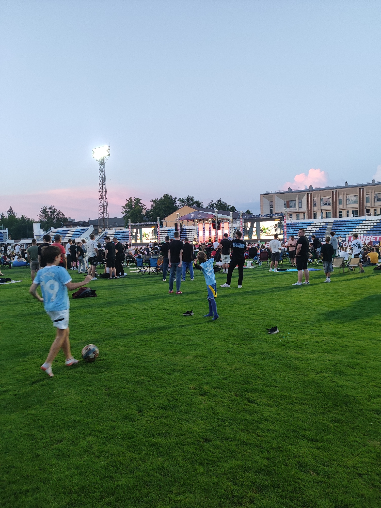 This image captures an outdoor gathering on a grassy field in front of a stadium in Brest, Belarus. The sky is a pale blue with hints of pink from the setting sun, and a tall stadium light tower illuminates the scene. A stage with a large screen is set up at the far end of the field, with a crowd of people gathered around it, some seated in chairs and others standing. In the foreground, two young boys are playing soccer on the grass. One boy, wearing a light blue jersey with the number "19" on the back, kicks the ball while the other, in a blue and yellow uniform, watches. The overall atmosphere appears relaxed and festive, with people enjoying an evening event outdoors.