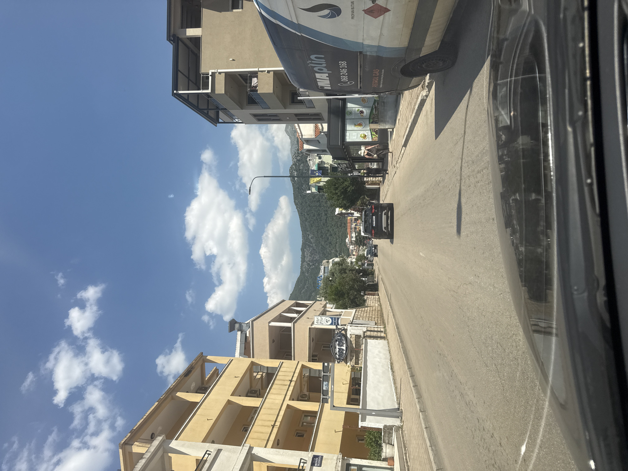 The image captures a street scene in Budva, Montenegro, on a sunny day with scattered clouds. The perspective is from inside a vehicle, looking forward. The street is paved with asphalt and has a sidewalk on the right.
On the left side of the street, there are several buildings, predominantly yellow and beige, with balconies and windows. One building has a sign that appears to say "Stari Ribar."
On the right side of the street, a large white van with dark blue and black accents is parked. The van has text on its side, including "INAPLIN" and a phone number. Further down the street, there are more buildings, including one with a modern awning and a business displaying food items.
Several cars are visible on the road, including a black SUV in the foreground and other vehicles further ahead. Two pedestrians are walking on the sidewalk on the right side of the street.
In the background, hills covered with greenery rise up, with more buildings scattered on their slopes. The sky is a bright blue with fluffy white clouds. The overall impression is of a typical urban environment in a Mediterranean setting.