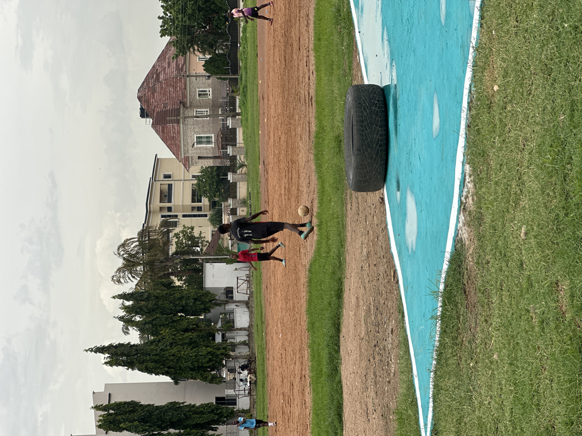 The image depicts an outdoor sports scene, likely a soccer field, in Abuja, Nigeria. Several individuals are present, engaged in or observing the activity. In the foreground, a person in a black jersey with the number 11 is kicking a soccer ball. Nearby, another person in a red jersey is also on the field, appearing to be involved in the game. Further in the background, a person in a pink top is running. Several other figures are visible in the distance, some appearing to be spectators or participants in a different area of the field.
The setting includes a dirt playing surface adjacent to a vibrant turquoise-colored area with white markings, possibly a court or a track. A large, worn tire rests on the edge of the turquoise area, likely used for training exercises. The surrounding environment features lush green grass, trees, and buildings in the background, indicating an urban or suburban location. The architecture of the buildings, with their distinct roofing and window styles, along with the lush vegetation, suggests a tropical or subtropical climate.
The sky is overcast with thick clouds, hinting at either a cloudy day or the potential for rain. The lighting is diffuse, suggesting it is not midday. The overall impression is one of a casual sporting event or training session taking place in a community setting. No legible text is visible on any of the subjects or objects.