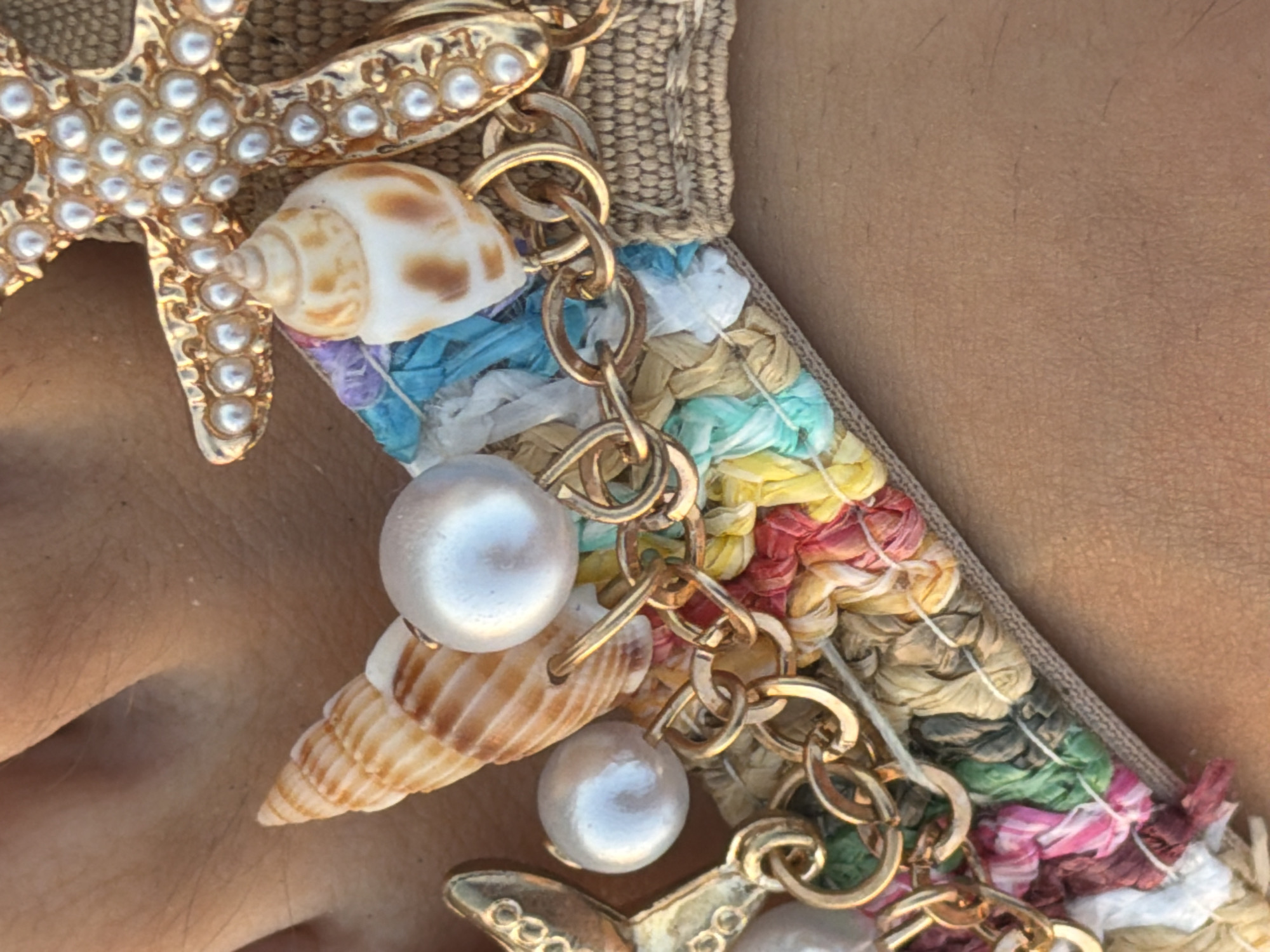 A close-up, overhead shot focuses on a decorative sandal strap with a nautical theme. The strap is woven with colorful raffia in shades of pink, yellow, blue, and white, interspersed with gold chain links. Various embellishments are attached, including two iridescent white pearls, a small brown and white striped seashell, and a larger, elongated seashell. A gold starfish charm adorned with small pearls is also visible, partially obscuring the strap. The background is a person's skin, likely an arm or leg, with subtle texture and fine hairs. The lighting suggests an outdoor setting, possibly during the day. The scene is located in Meneou, Cyprus.