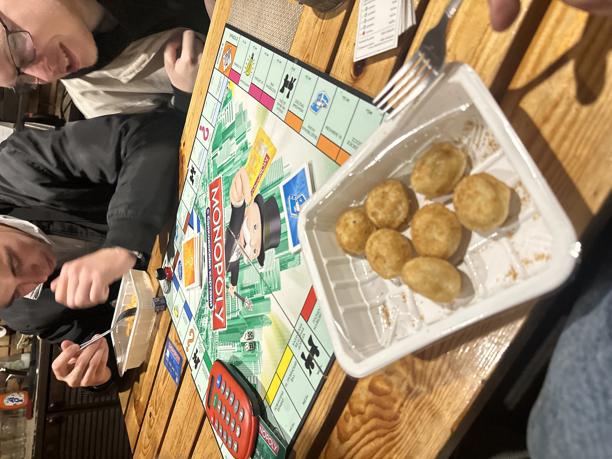 The image depicts a group of young adults playing a game of Monopoly. The game board is centrally located on a wooden table. Several Monopoly cards, dice, and a red calculator are visible on the board. One individual, seen from the chest up, is actively engaged in the game, holding a fork and interacting with a white plastic container of food placed on the table. The container holds several small, golden-brown, spherical food items. Another individual, wearing glasses, is partially visible to the right, also appearing to be participating in the game. The background suggests an indoor setting, possibly a home or a casual dining area, with a glimpse of a brick wall and kitchen cabinets. The overall atmosphere is one of casual social interaction and shared activity. The image does not provide any specific information that would allow for its location to be identified as "Востец Снт, Russia."