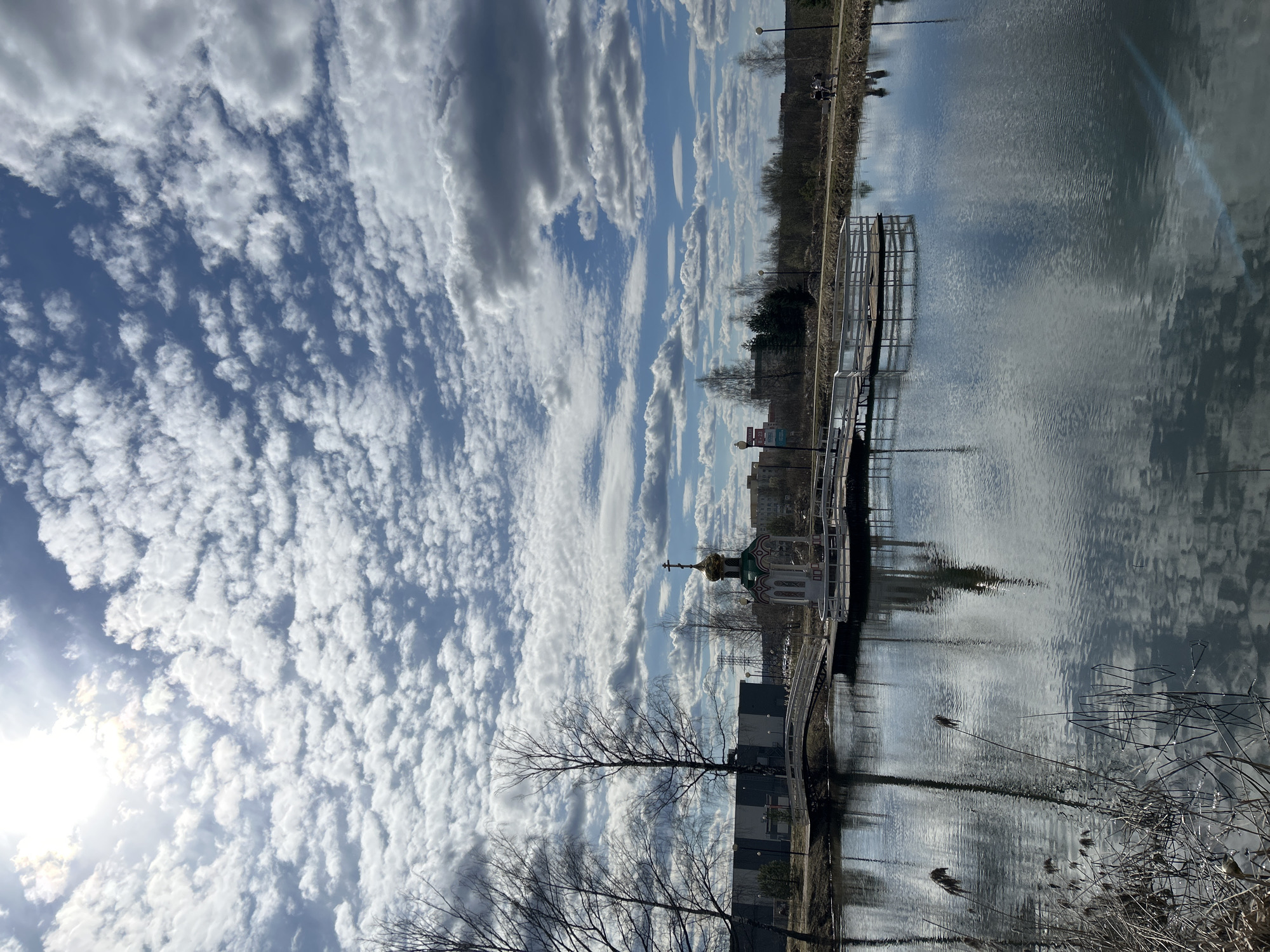 The image is a landscape oriented scene with a body of water in the center. The sky is filled with altocumulus clouds. There is a walkway leading to a small structure with a green and gold dome, possibly a chapel. This is located at the city of Klin, Russia. There are trees and reeds near the edge of the water. Some buildings are visible in the background. A walkway goes behind the chapel and is reflected in the water.