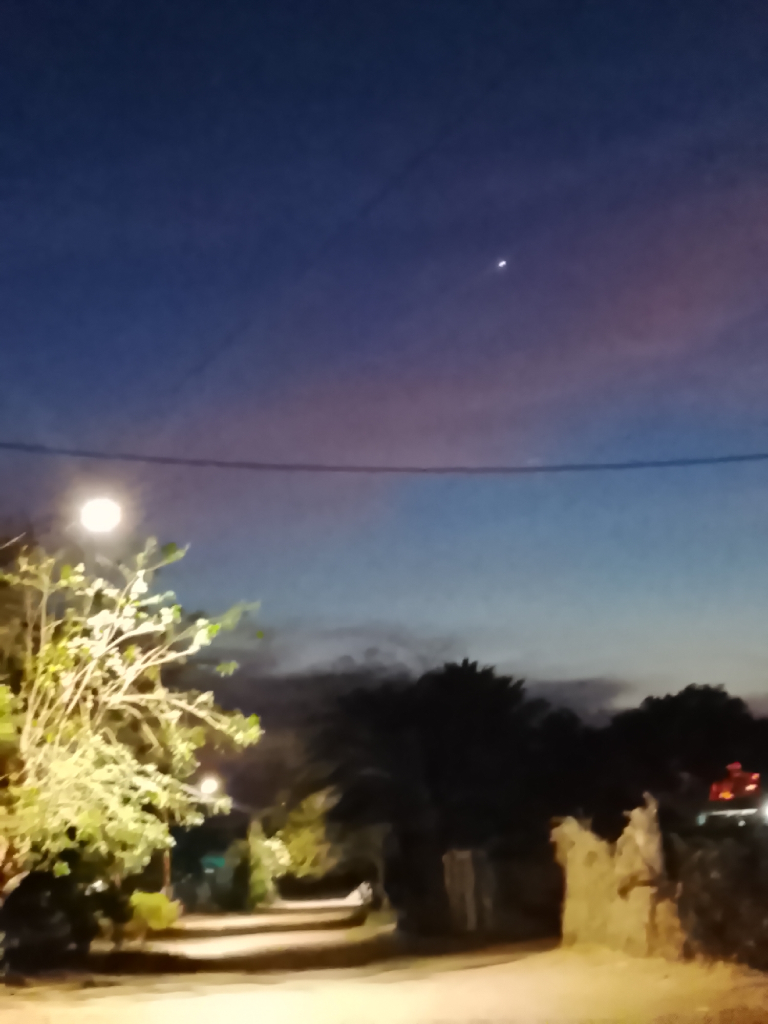 This low-light image captures a quiet street or path, likely at dusk or early night, in Altagracia, Venezuela. The sky features a deep blue overhead, transitioning to purple and faint reddish-orange towards the horizon, with subtle cloud formations. A single, bright celestial object, possibly a star or planet, shines prominently in the upper right portion of the sky. A dark power line or wire stretches horizontally across the frame.
On the left side, a lush tree or large bush with green foliage is brightly illuminated by a nearby streetlight, casting a warm, yellowish glow. The light source itself appears as a bright, circular orb. The road, possibly unpaved, is dimly lit and recedes into the distance, bordered by silhouetted trees and vegetation. On the right, a low, light-colored stone wall or structure runs alongside the path. In the far distance on the right, blurry red and orange lights suggest a vehicle or building. The scene conveys a peaceful, solitary atmosphere with no visible people or specific activities. The image has a soft focus, contributing to its tranquil mood. No visible text is present.