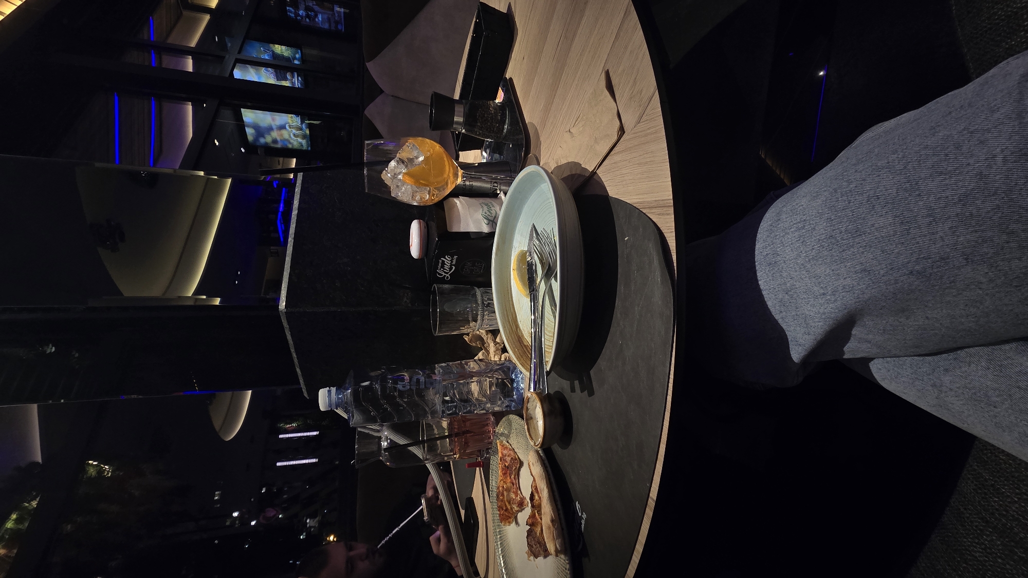 The image shows a round table in a dimly lit setting, possibly a restaurant or bar. On the table, there is a variety of items including a drink with ice and an orange slice, a bottle labeled "Linda," plates with remnants of food, a water bottle, and utensils. A person wearing jeans is seated at the table. The background suggests an urban environment, with lights and structures visible. The location is possibly in Stara Zagora, Bulgaria.