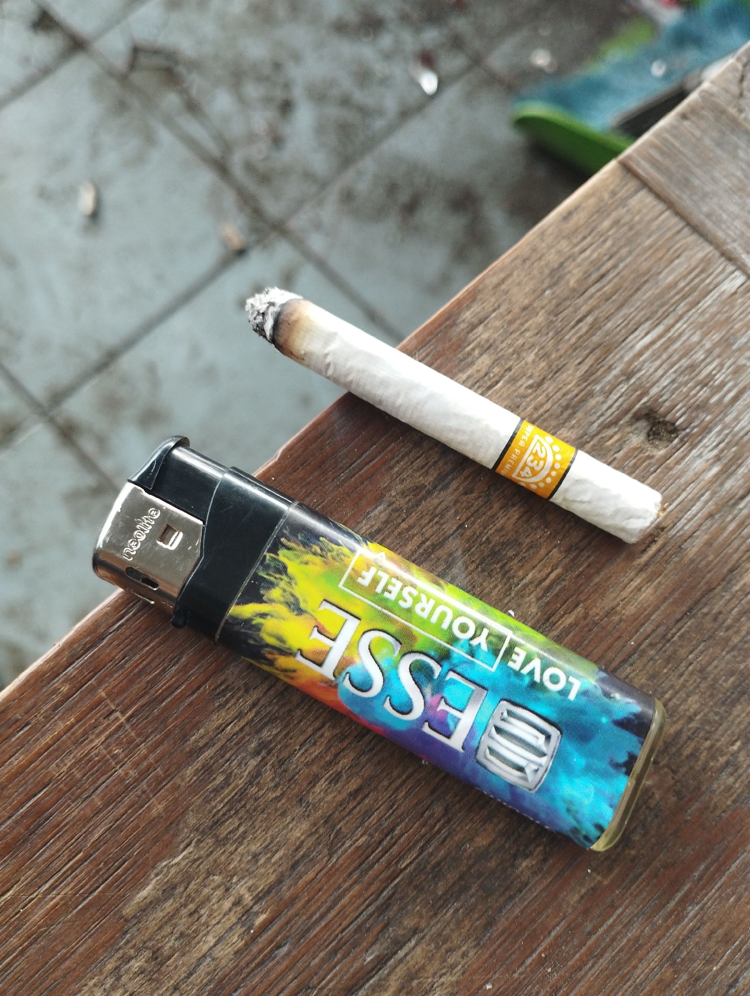 A lit cigarette and a lighter are on a wooden surface. The cigarette has a yellow band around it with the number "234". The lighter has the word "ESSE" and the phrase "LOVE YOURSELF" on it. The location appears to be outdoors, possibly near a tiled surface. There is no indication of the city Lhokseumawe, Indonesia in the image.
