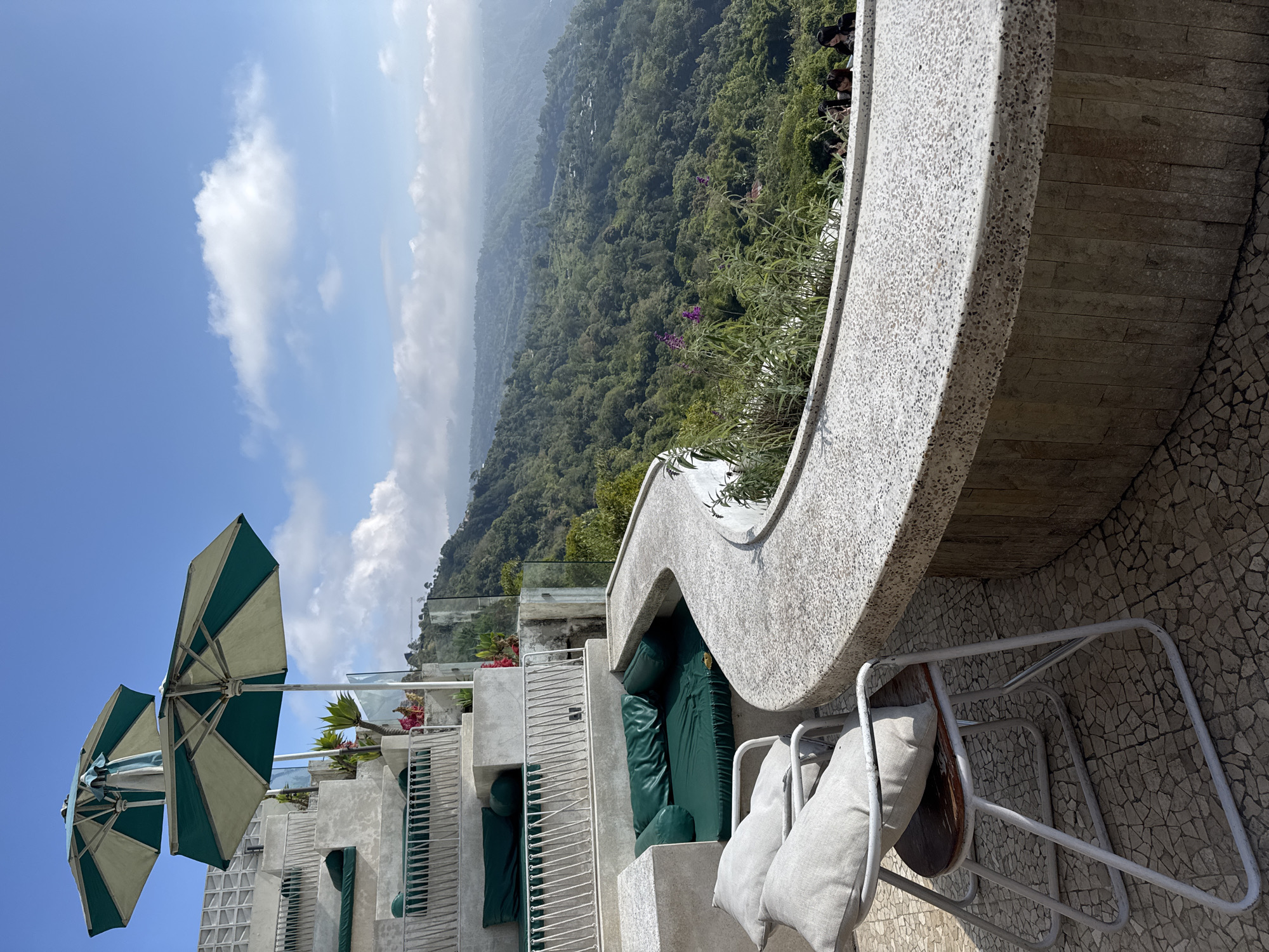 The image depicts an outdoor seating area overlooking a lush, mountainous landscape in Kintamani, Indonesia. In the foreground, two light-colored chairs with cushions are positioned on a patterned tile floor. Adjacent to the chairs is a curved, stone counter with a speckled surface, supported by a stone-clad base. Behind the seating area, a tiered, concrete structure features built-in green cushioned seating areas. Two teal and white umbrellas are positioned above this tiered seating. The background is dominated by a verdant mountain range covered in dense forest, with scattered buildings visible amongst the trees. The sky is blue with scattered white clouds. A few individuals are visible near the counter, looking out at the view.