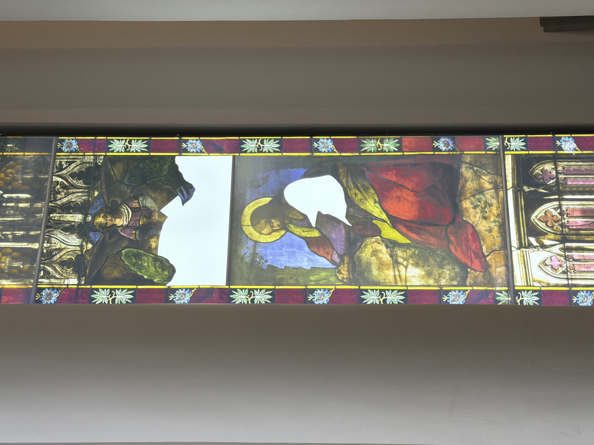 The image shows a stained glass window, possibly from a church or cathedral. The window depicts religious figures, with the central figure appearing to be kneeling, adorned with a halo. The colors are rich and include reds, greens, blues, and golds. Parts of the stained glass are missing, creating stark white gaps in the imagery. The location cannot be determined from this single image. There are no visible people present besides the depictions in the stained glass. The window has a border design of flowers and foliage. The window is installed at a certain height up on a wall.