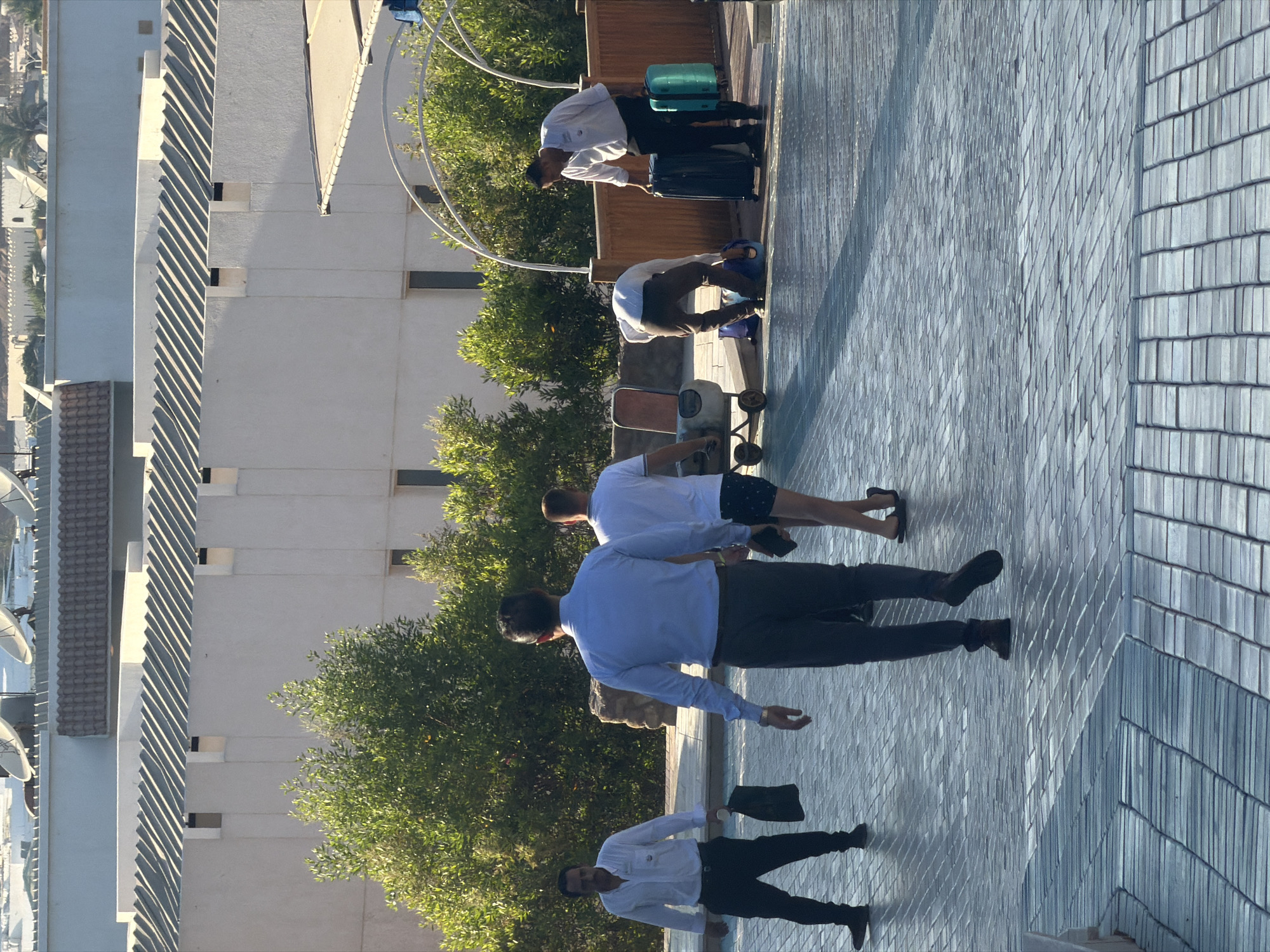 The image depicts an outdoor scene, likely in a resort or hotel setting in Sharm El Sheikh, Egypt. In the foreground, a paved pathway with a patterned stone surface is visible, reflecting sunlight. Several individuals are present.
On the left, a man in a white collared shirt and black pants is walking, holding a white cup and a black bag. To his right, a man in a light blue dress shirt and dark pants is walking away from the camera. Behind him, a man in a white t-shirt and patterned shorts is also walking away.
Further into the scene, another man in a white shirt is bending down, interacting with a blue bag. Next to him, a man in a white collared shirt and black pants is standing, holding a dark suitcase in his right hand and a turquoise suitcase in his left hand. A luggage cart with an orange seat is also visible.
The background features lush green trees and buildings with white walls and sloped roofs. The overall ambiance suggests a warm, sunny day, possibly during a time of arrivals or departures at a travel destination.