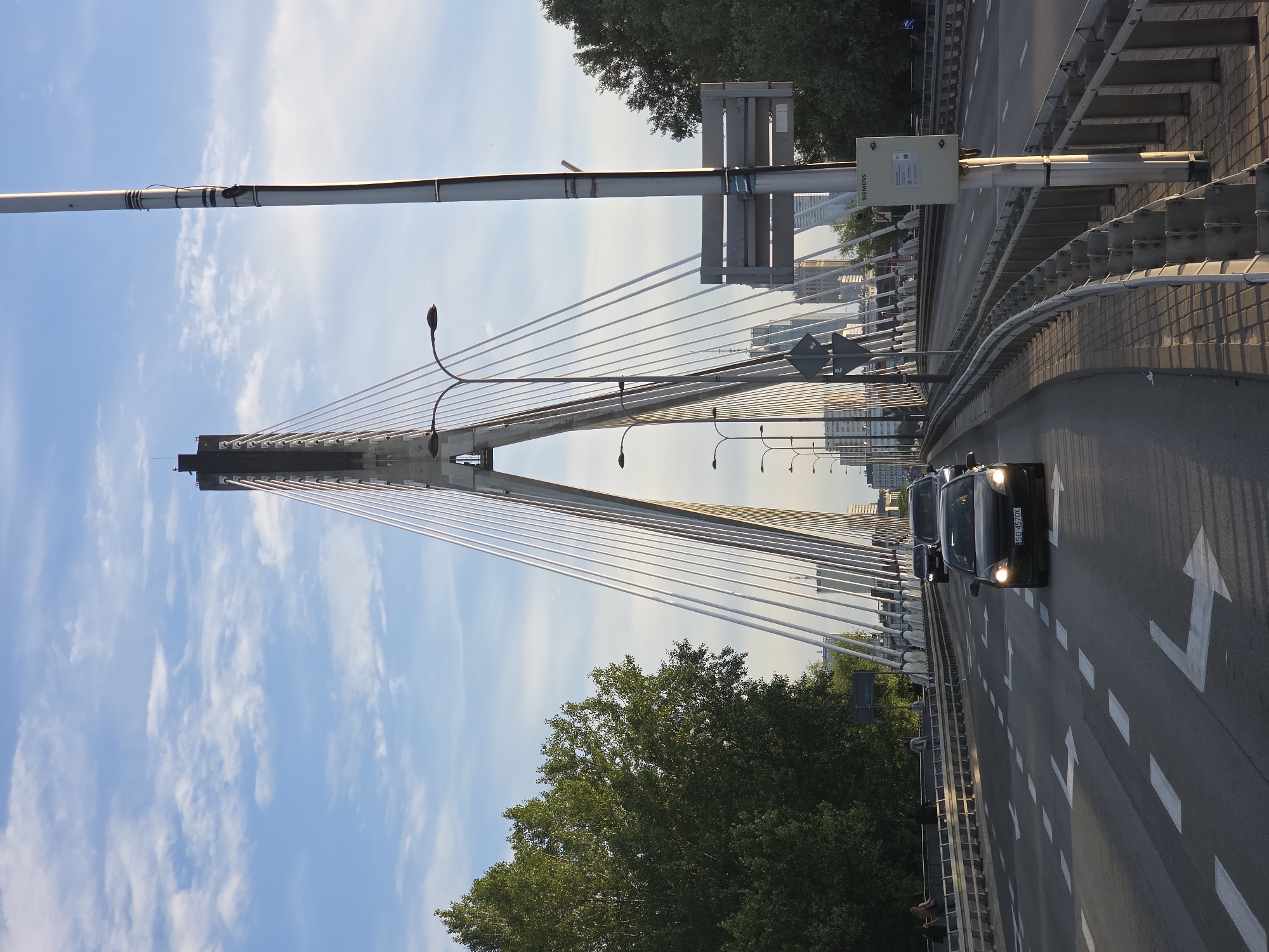 The image shows the Świętokrzyski Bridge in Warsaw, Poland. It is a cable-stayed bridge that spans the Vistula River. The bridge is made of concrete and steel, and it is supported by two towers. The towers are 66 meters high, and the bridge deck is 404 meters long. The bridge was built between 1997 and 2000. The image shows traffic on the bridge, including cars and buses. There are also trees and buildings in the background. The sky is blue with some clouds.