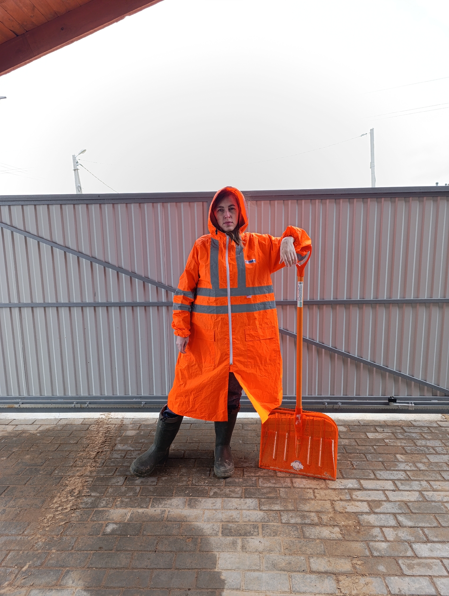 A woman in an orange raincoat stands in front of a gray metal fence, leaning on an orange snow shovel. She wears black rubber boots. The ground is paved with gray bricks. The scene is in Chaikovsky, Russia.