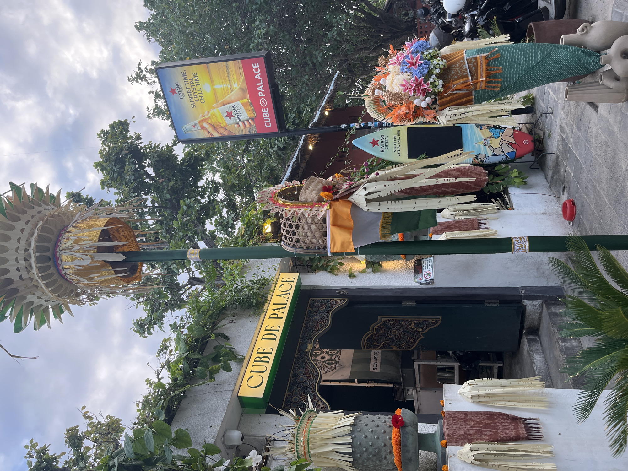 This is a street-level, eye-level, medium shot of an outdoor area in Denpasar, Indonesia, likely in front of a business. In the center, a dark green door with ornate carvings is visible, leading into what appears to be a shop or restaurant. Above the door, a rectangular green sign with yellow lettering reads "CUBE DE PALACE." To the left of the entrance, a large, grey planter with orange and red flowers sits on a pedestal. The facade of the building is white and covered in lush green vines.
To the right of the door, a tall green pole stands, adorned with traditional Balinese decorations, including woven baskets and fabric streamers. A large, vertical advertisement for "Bintang Crystal" beer is mounted on this pole, featuring an image of a sunset and a bottle of beer, with text that reads "SUNSET TIME, CRYSTAL CHILL TIME." Further to the right, a colorful floral arrangement is prominently displayed, draped in patterned fabric.
The ground is paved with stone tiles. In the background, trees with dense foliage can be seen, and a cloudy sky is visible overhead. Several motorcycles are parked to the far right of the frame, suggesting a busy street or entertainment area. The overall impression is one of vibrant cultural decoration and a commercial establishment. There are no people clearly visible in the image.