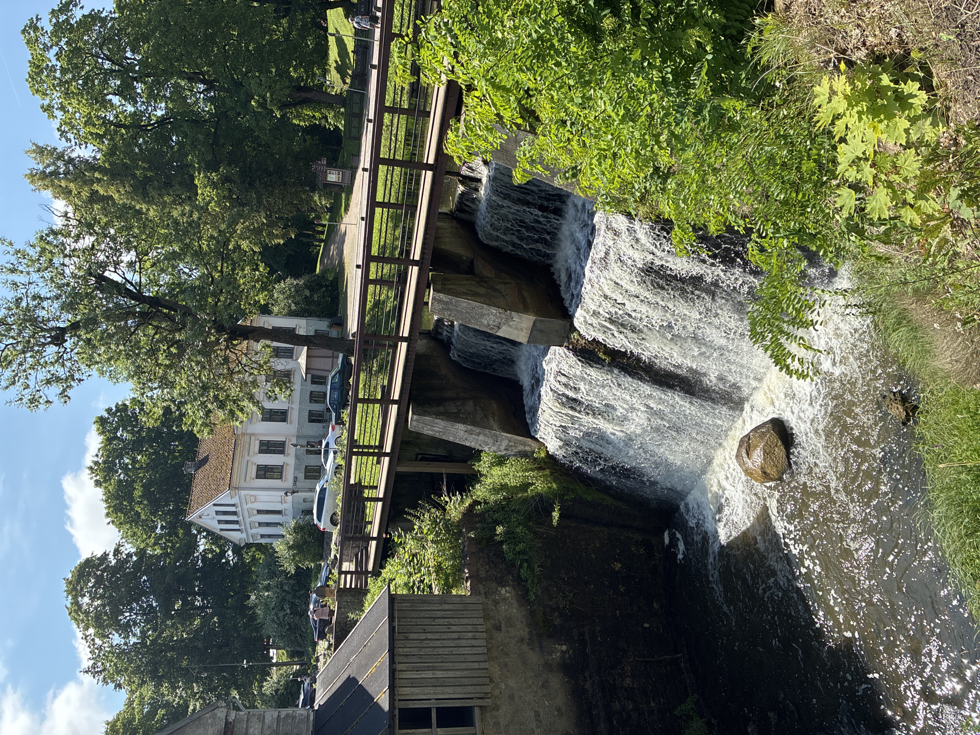 The image depicts a picturesque scene in Kuldīga, Latvia, likely near the Venta River. A man-made waterfall cascades over a concrete structure, with water flowing into a dark pool below. A large, rounded boulder rests in the foreground of the pool, partially submerged in the turbulent water. A wooden pedestrian bridge spans across the top of the waterfall, supported by concrete pillars. On the left side of the bridge, a small, rustic wooden building with a dark, sloped roof is visible. In the background, a white, multi-story building with a prominent red-tiled roof stands amidst lush green trees. Several cars are parked on a road in front of the white building, and a few individuals are seen walking on the bridge and near the cars. The environment is characterized by abundant greenery, indicating a summer or spring setting. The sky is blue with scattered white clouds.