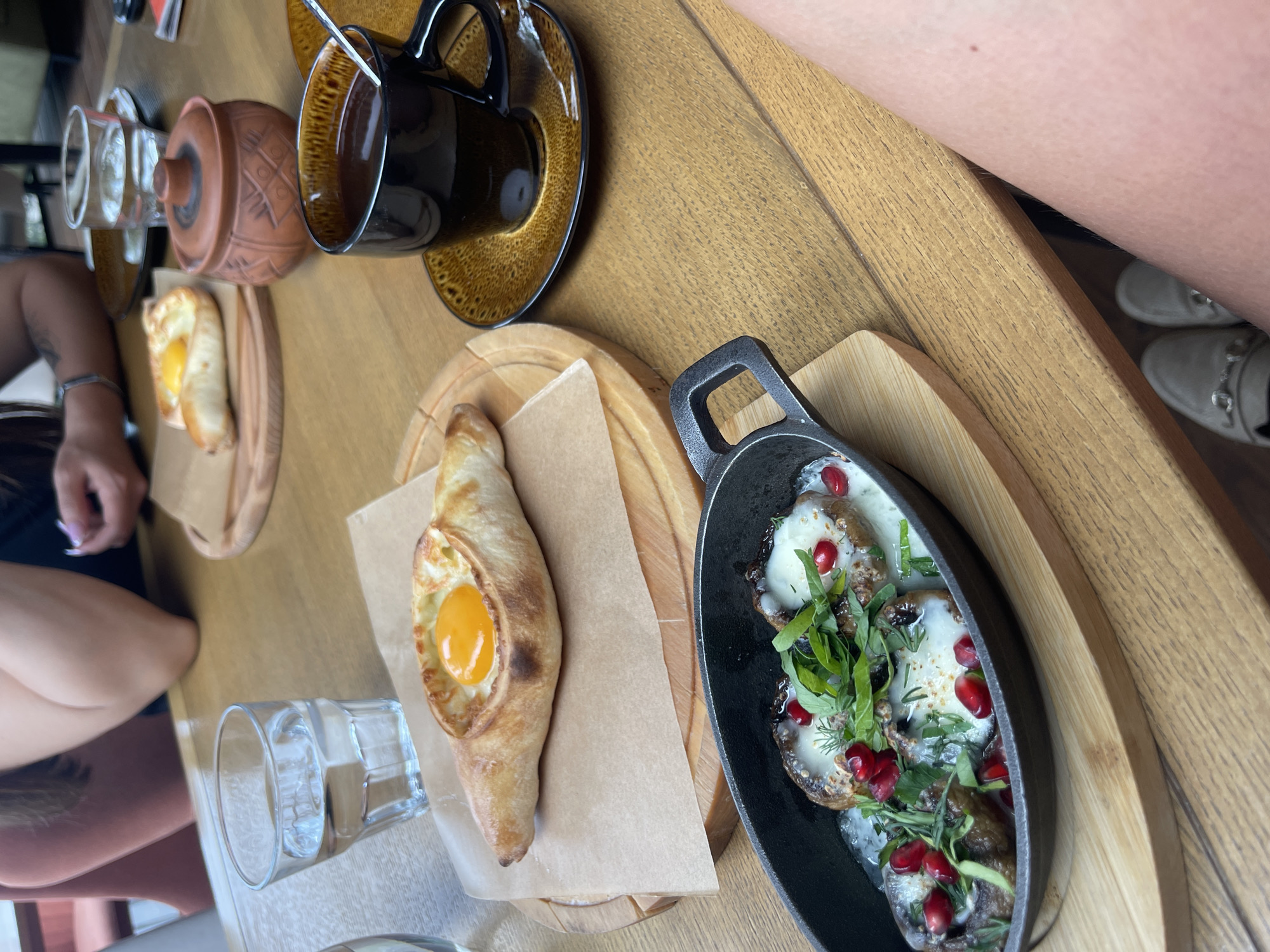 The image captures a restaurant table in Perm, Russia, displaying a culinary scene. A cup of coffee sits atop a saucer, alongside a ceramic jar and a small plate with khachapuri. Adjacent to this, another plate holds an elongated khachapuri with an egg yolk at its center. The composition is completed by a cast-iron skillet containing a dish garnished with greens and pomegranate seeds. To the left, a person's arm and hand are visible, while on the right, a partial view of a person's leg and shoe can be seen. The table itself is made of wood.