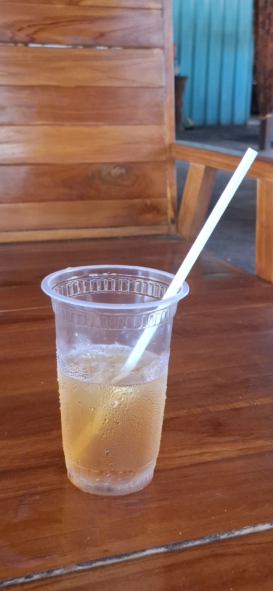 In Desa Sobokerto, Indonesia, a close-up shot captures a plastic cup filled with amber liquid and ice. The cup's exterior is beaded with condensation, indicating a cold beverage. A white straw extends from the liquid upwards, at an angle, and exits the cup near its rim. The cup rests on a polished wooden table, the surface of which is smooth and shows faint grain patterns. In the background, a wooden chair with horizontal slats is visible, suggesting an outdoor or semi-outdoor seating area. Beyond the chair, a blue corrugated metal wall forms the backdrop. The lighting appears to be natural, casting soft shadows and highlighting the condensation on the cup.