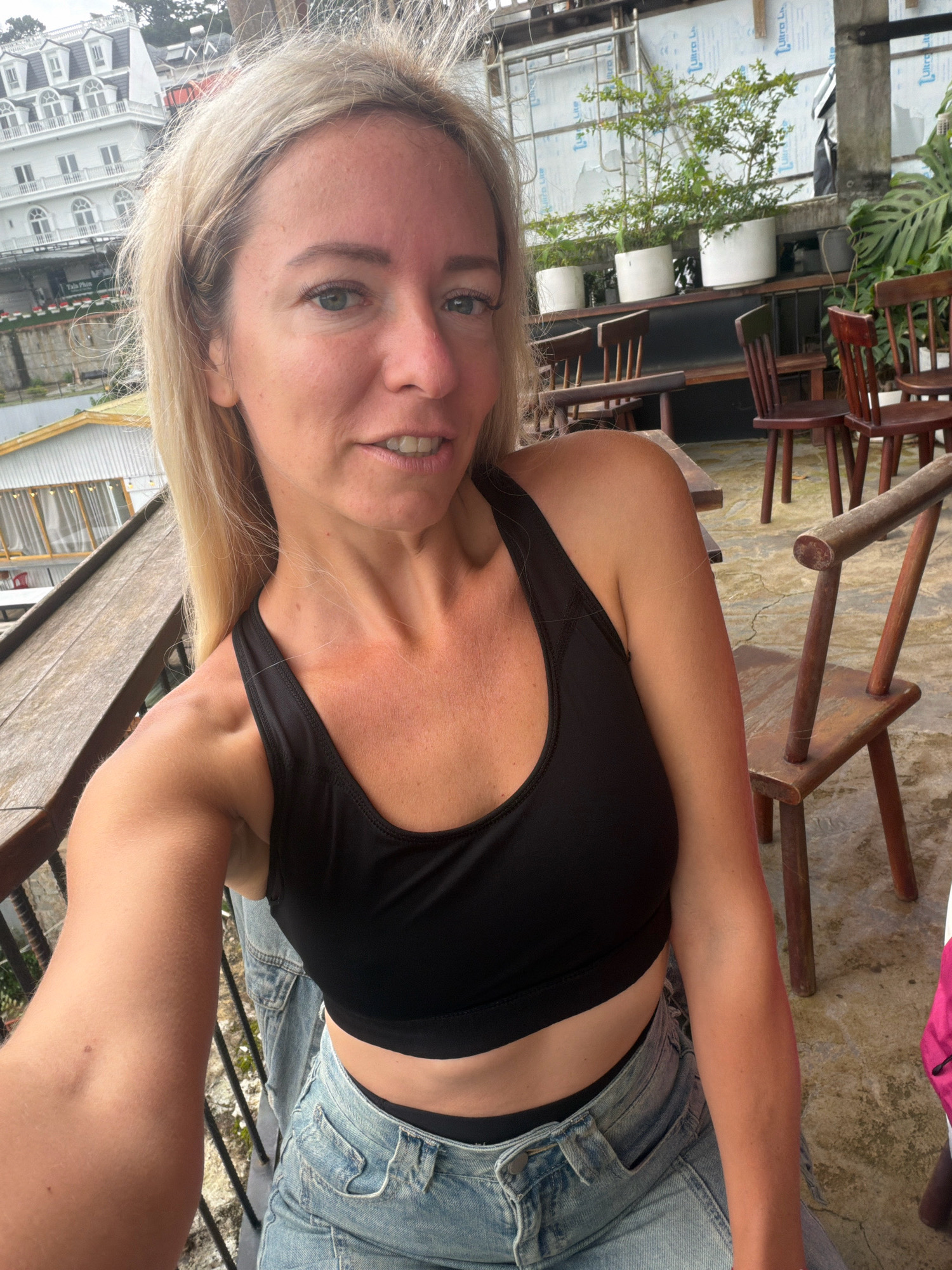 A woman with blonde hair, wearing a black sports bra and denim jeans, is captured in a selfie. She is standing on a wooden balcony, with a rustic wooden chair to her right and a railing to her left. In the background, a white building with a distinct French colonial architectural style is visible, its white facade contrasting with the grey sky. Beyond the building, lush green hills suggest a mountainous landscape. A glimpse of plant pots and more wooden furniture can be seen behind her, implying an outdoor seating area at a cafe or restaurant. The overall ambiance points to a location in Da Lat, Vietnam, known for its French colonial architecture and elevated terrain.
